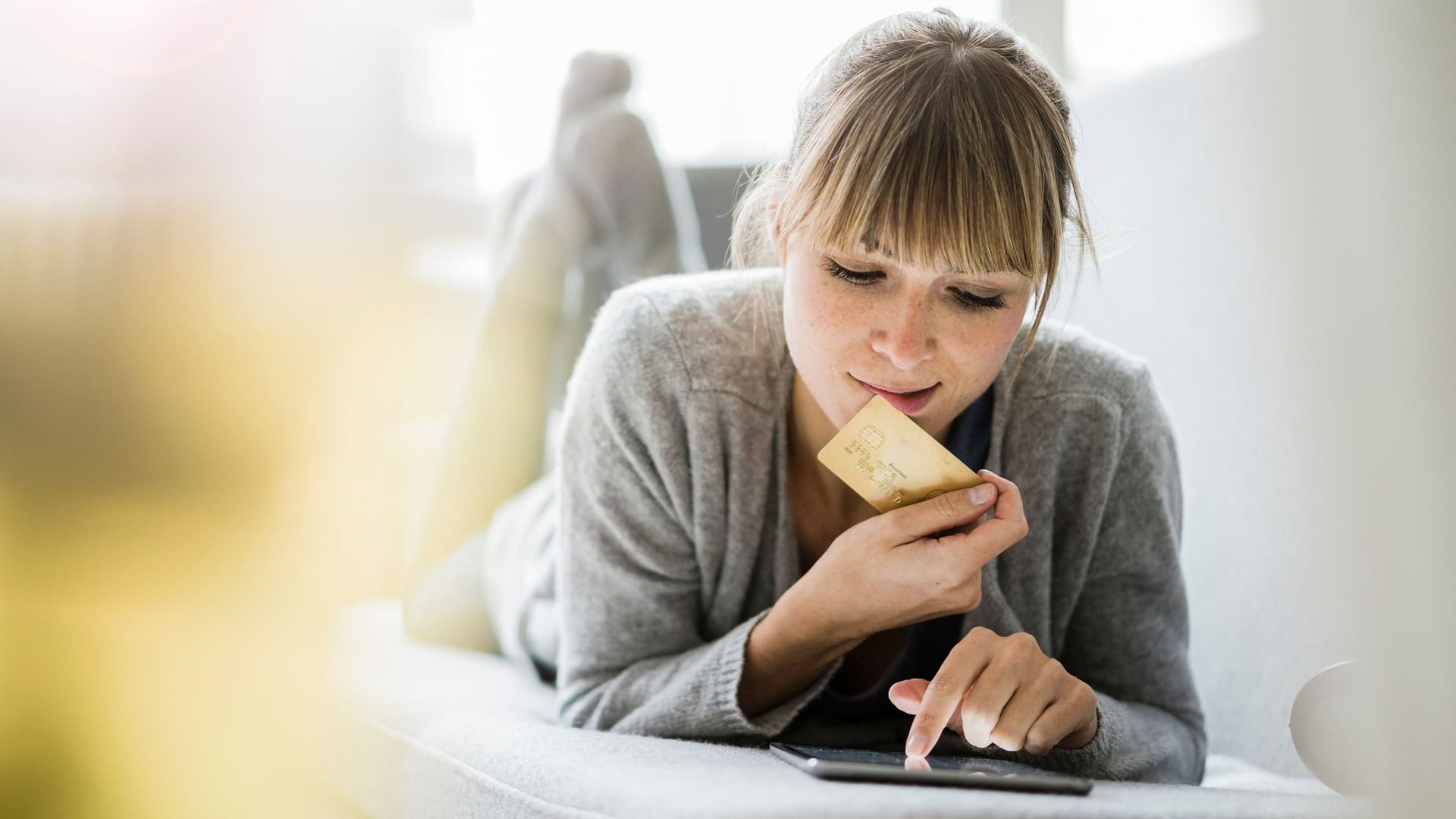 Junge Frau liegt mit ihrem Tablet auf einem Sofa, dabei hält sie eine Kreditkarte in ihrer rechten Hand.