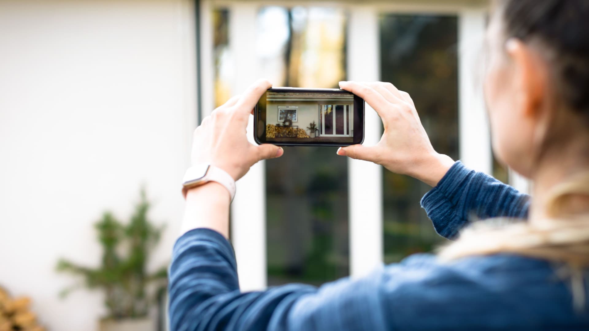 Eine Frau fotografiert mit ihrem Smartphone die Terrassentür eines Hauses von außen. Das Foto wird dabei auf dem Smartphone-Bildschirm angezeigt.