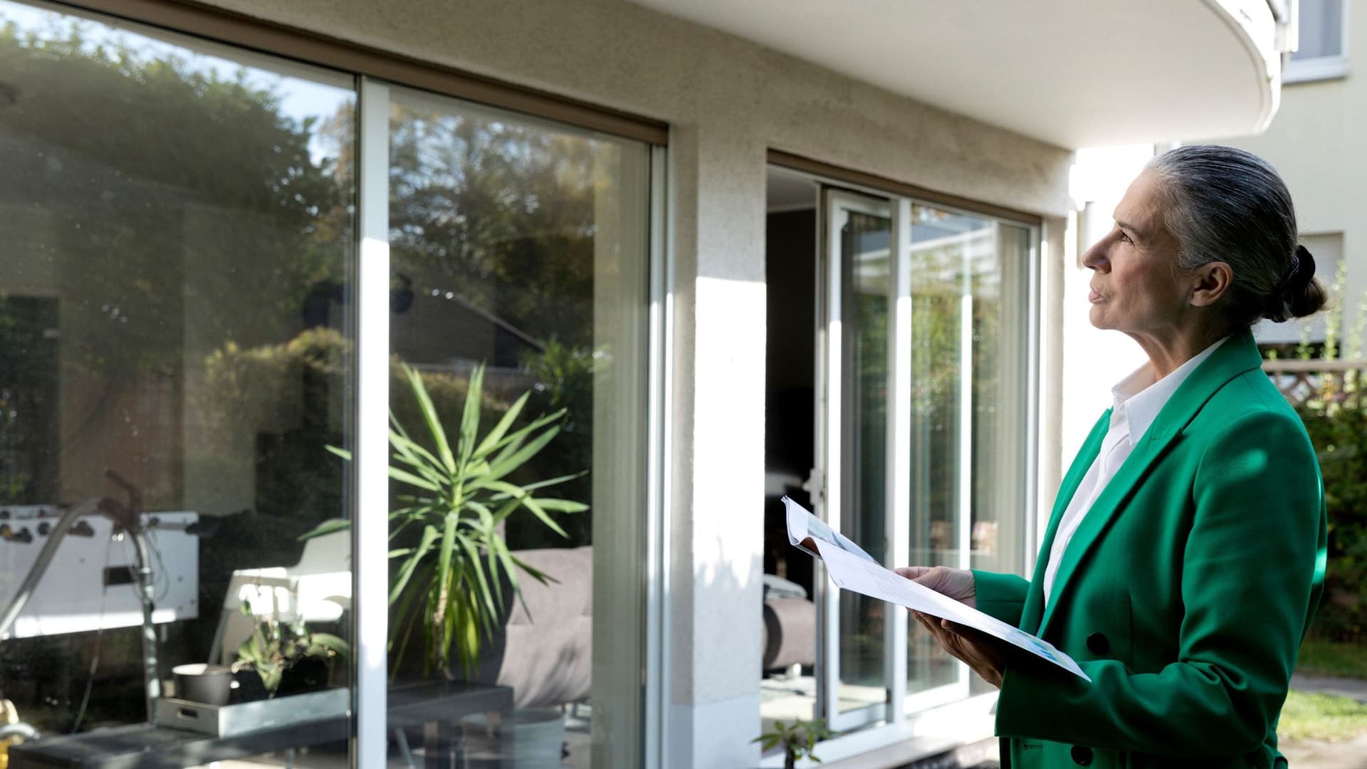 Eine Frau im grünen Blazer steht nachdenklich vor einem modernen Haus mit großen Glasfronten. In den Händen hält sie eine Broschüre oder Unterlagen.