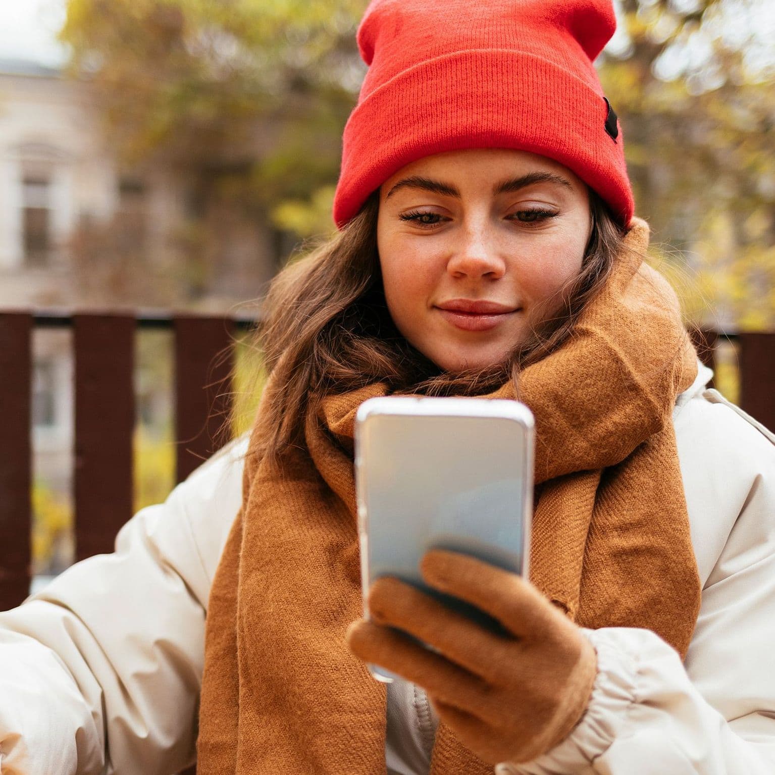 Eine Frau mit Mütze und Kaffee ind er Hand schaut auf ihr Smartphone