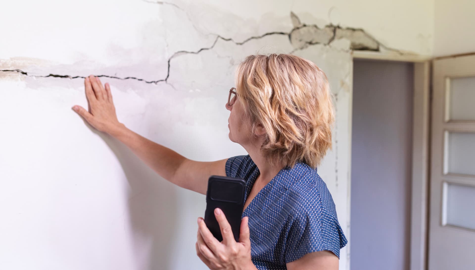 Frau betrachtet Riss in der Wand und hält ein Smartphone in der Hand