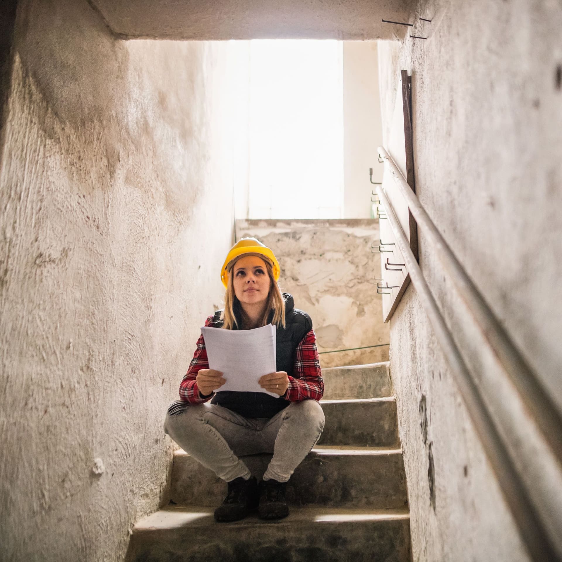 Junge Frau mit einem gelben Helm sitzt auf einer Steintreppe auf einer Baustelle. Sie hält Pläne in den Händen.