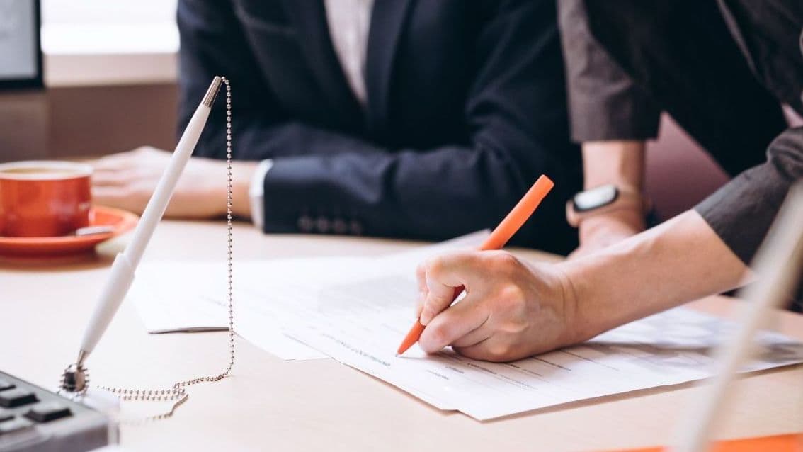 Eine Frau unterschreibt mit einem orangenen Stift ein Dokument auf einem Tisch, daneben sitzt eine weitere Person im Anzug. Auf dem Tisch liegen Unterlagen und eine Kaffeetasse. Die Szene vermittelt Vertragsabschluss oder Beratungsgespräch.