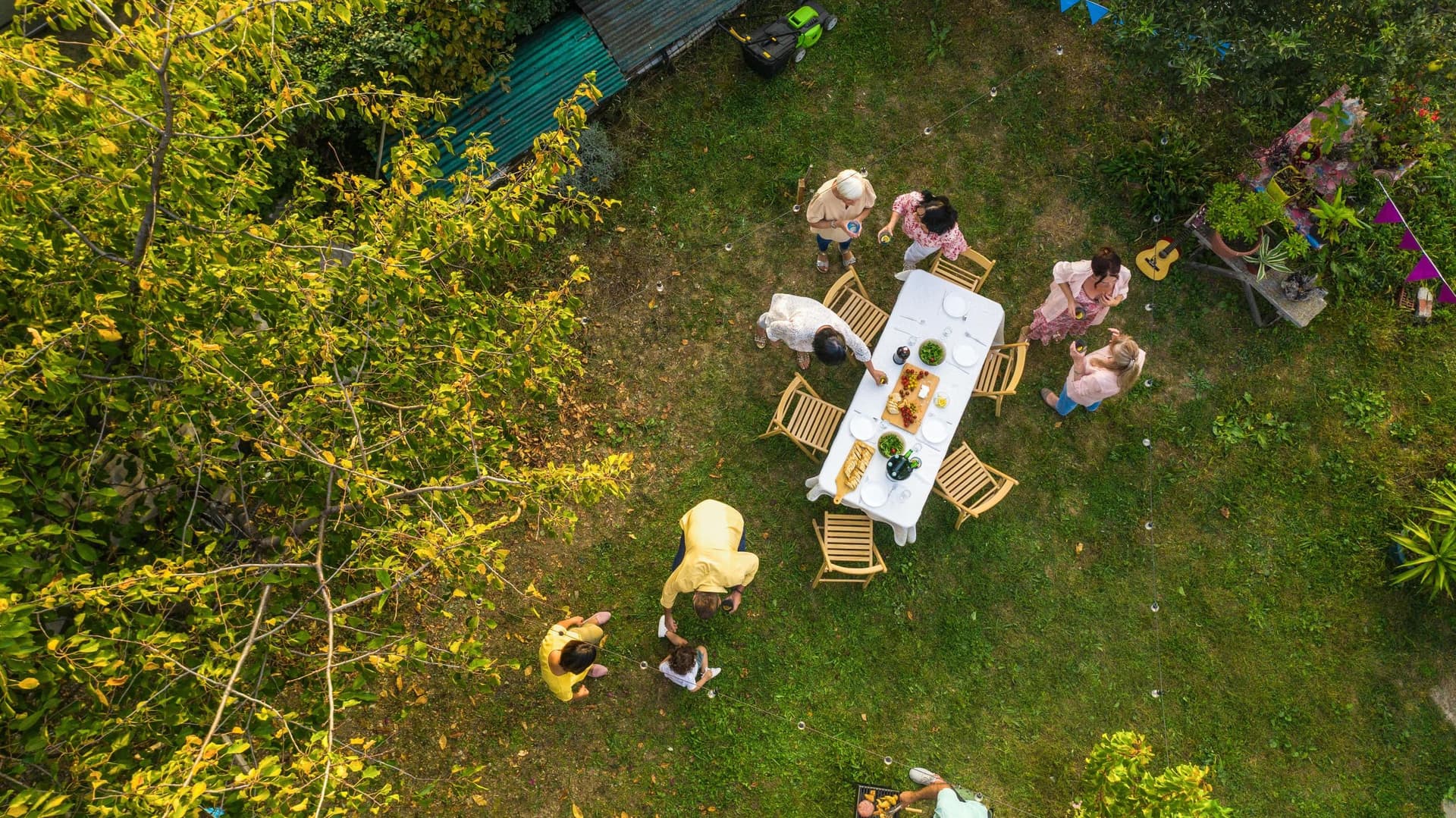 Drohnenaufnahme einer Gartenfeier: Mehrere Menschen sitzen und stehen im grünen Garten um einen gedeckten Tisch. Zwischen Bäumen und Pflanzen hängen bunte Wimpel und Lichterketten; im Hintergrund stehen eine Sitzbank und ein Rasenmäher.