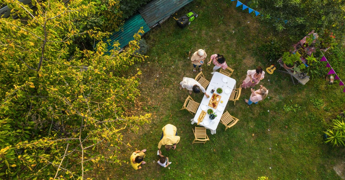 Drohnenaufnahme einer Gartenfeier: Mehrere Menschen sitzen und stehen im grünen Garten um einen gedeckten Tisch. Zwischen Bäumen und Pflanzen hängen bunte Wimpel und Lichterketten; im Hintergrund stehen eine Sitzbank und ein Rasenmäher.