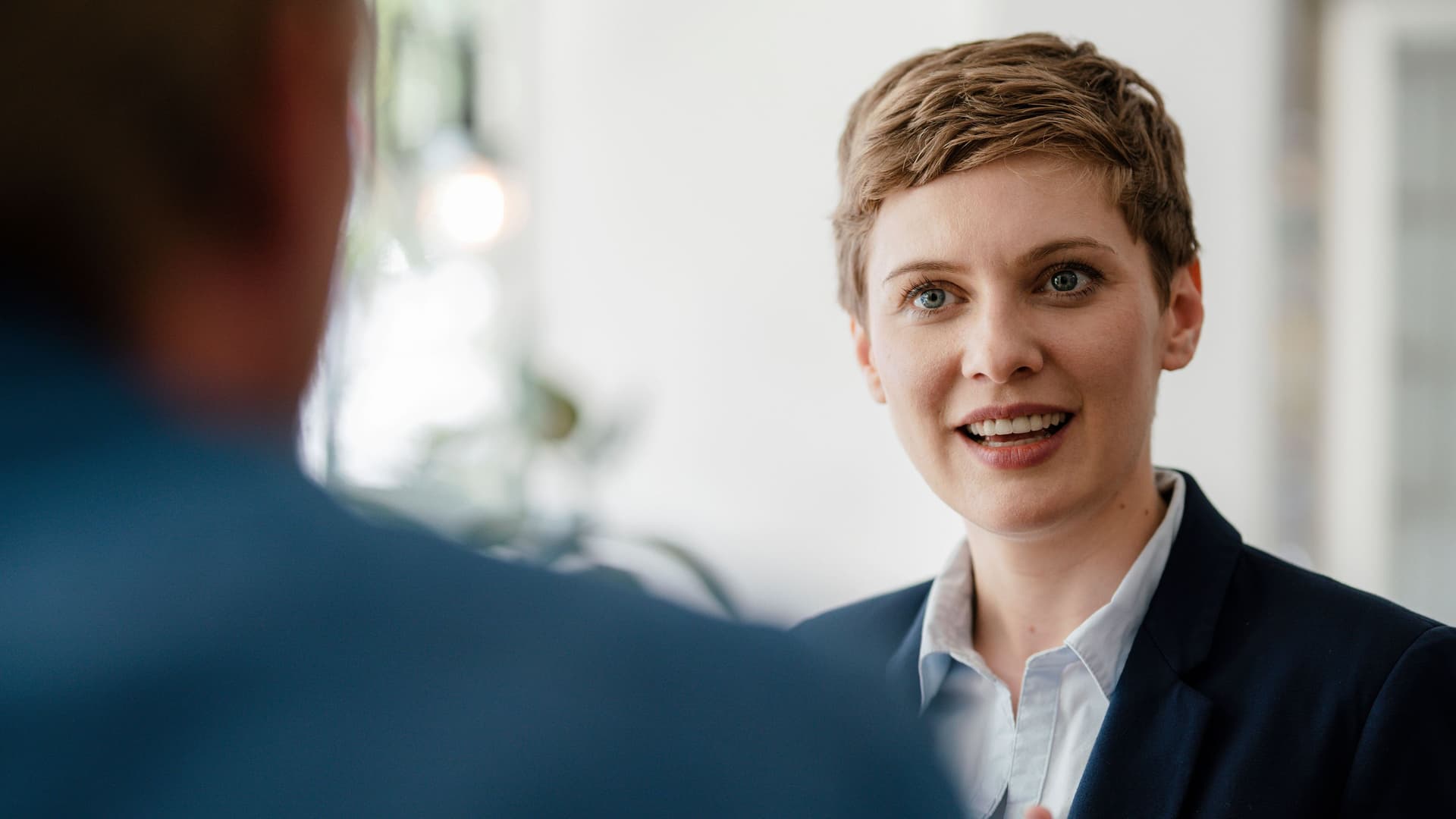 Porträt einer Frau in Business-Kleidung, die sich im Gespräch mit einem Kollegen befindet und mit den Händen dabei gestikuliert.