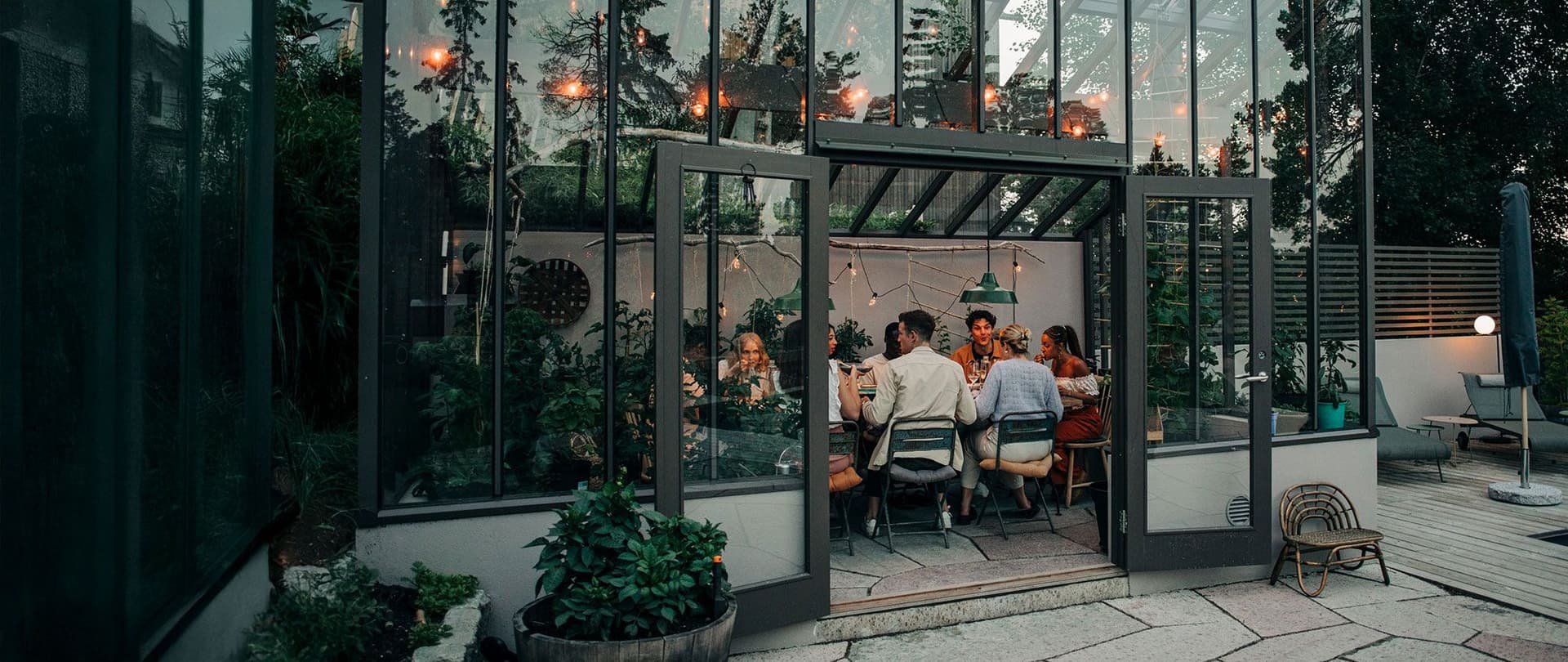 Gruppe von Menschen beim Abendessen in einem Wintergarten mit Pflanzen und Lichterketten