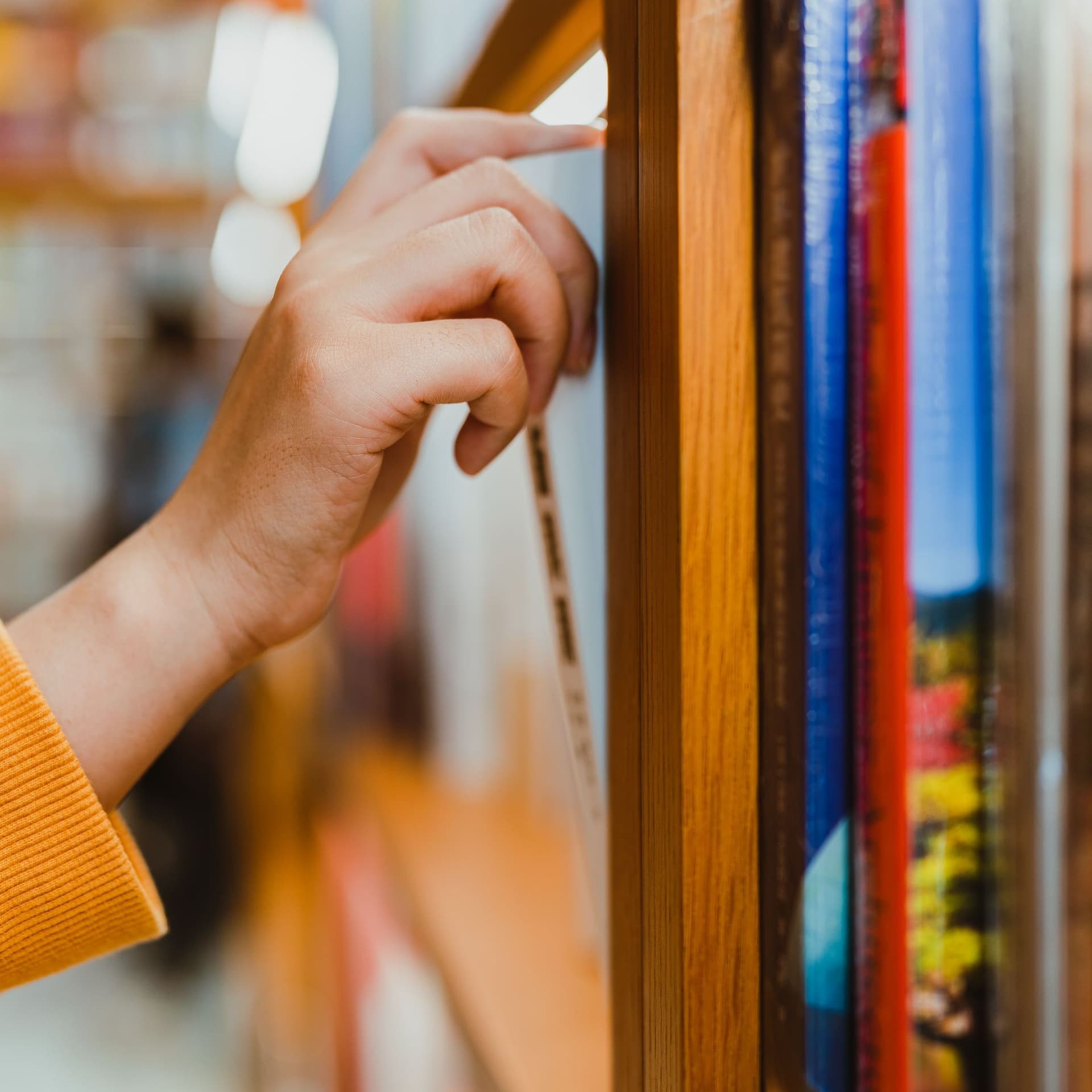 Seitliche Nahaufnahme von einem Arm mit orangefarbenen Pullover. Die Hand zieht ein Buch aus einem Bücherregal.