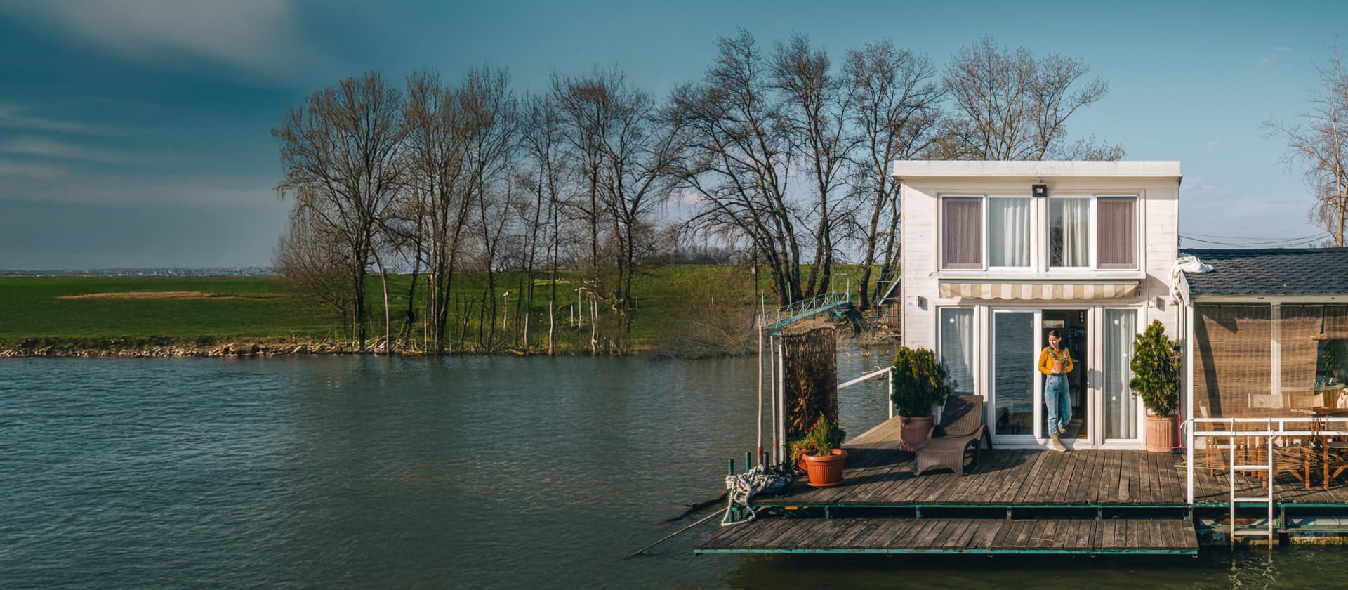 Weißes zweistöckiges Hausboot mit Holzterrasse, Pflanzenkübeln und überdachter Sitzecke auf einem Fluss bei Sonnenschein.