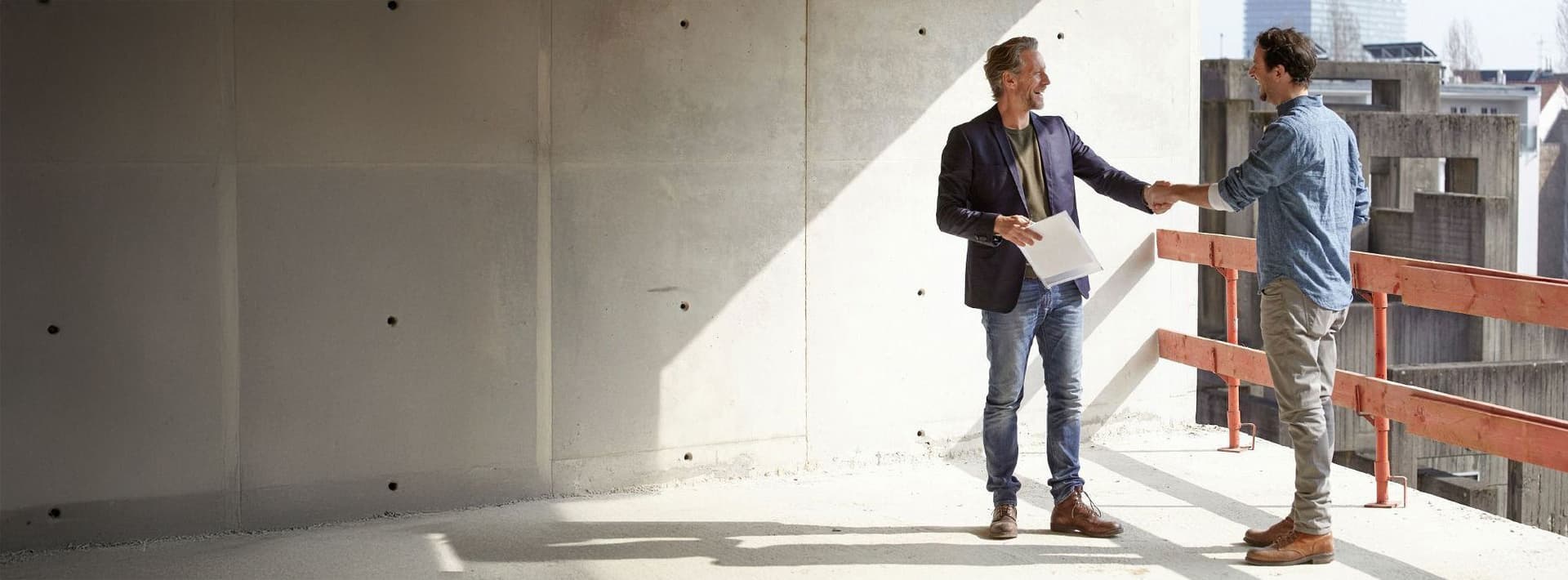 Zwei Männer stehen auf einer Baustelle und schütteln sich die Hand, einer hält Unterlagen, im Hintergrund Betonwände und Gerüstteile