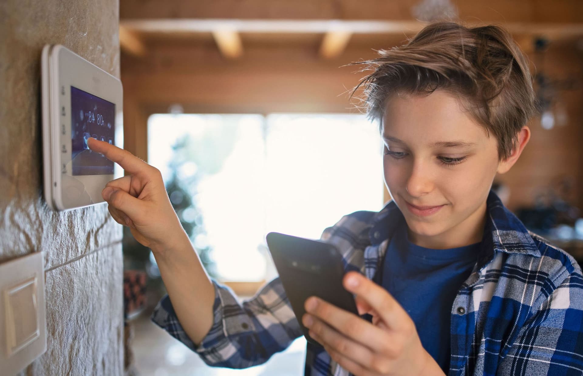 Junge Person bedient digitales Thermostat an Wand und schaut auf Smartphone in Wohnraum