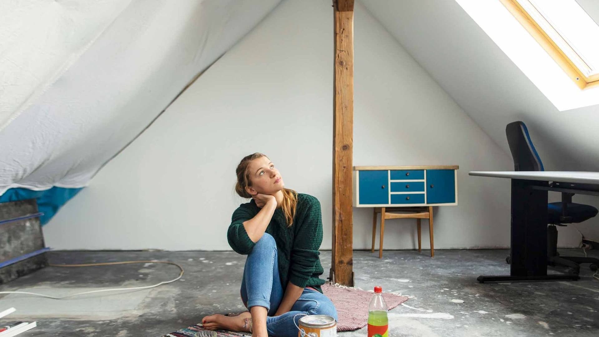 Junge Frau sitzt nachdenklich auf dem Boden eines unfertigen Dachgeschosses mit Farbeimer und Möbeln im Hintergrund