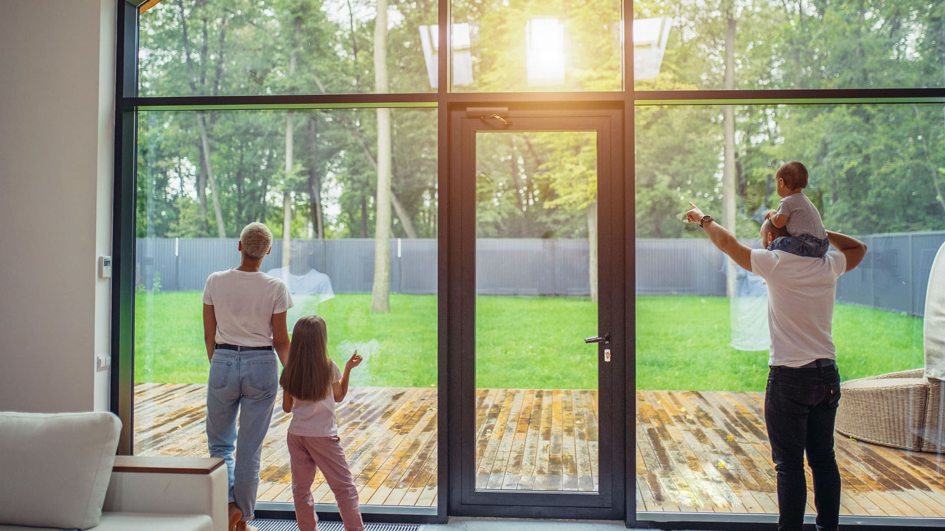 Familie (Eltern und zwei Kinder) steht im Wohnzimmer und blickt durch große Glasfenster auf eine Terrasse und einen grünen Garten.