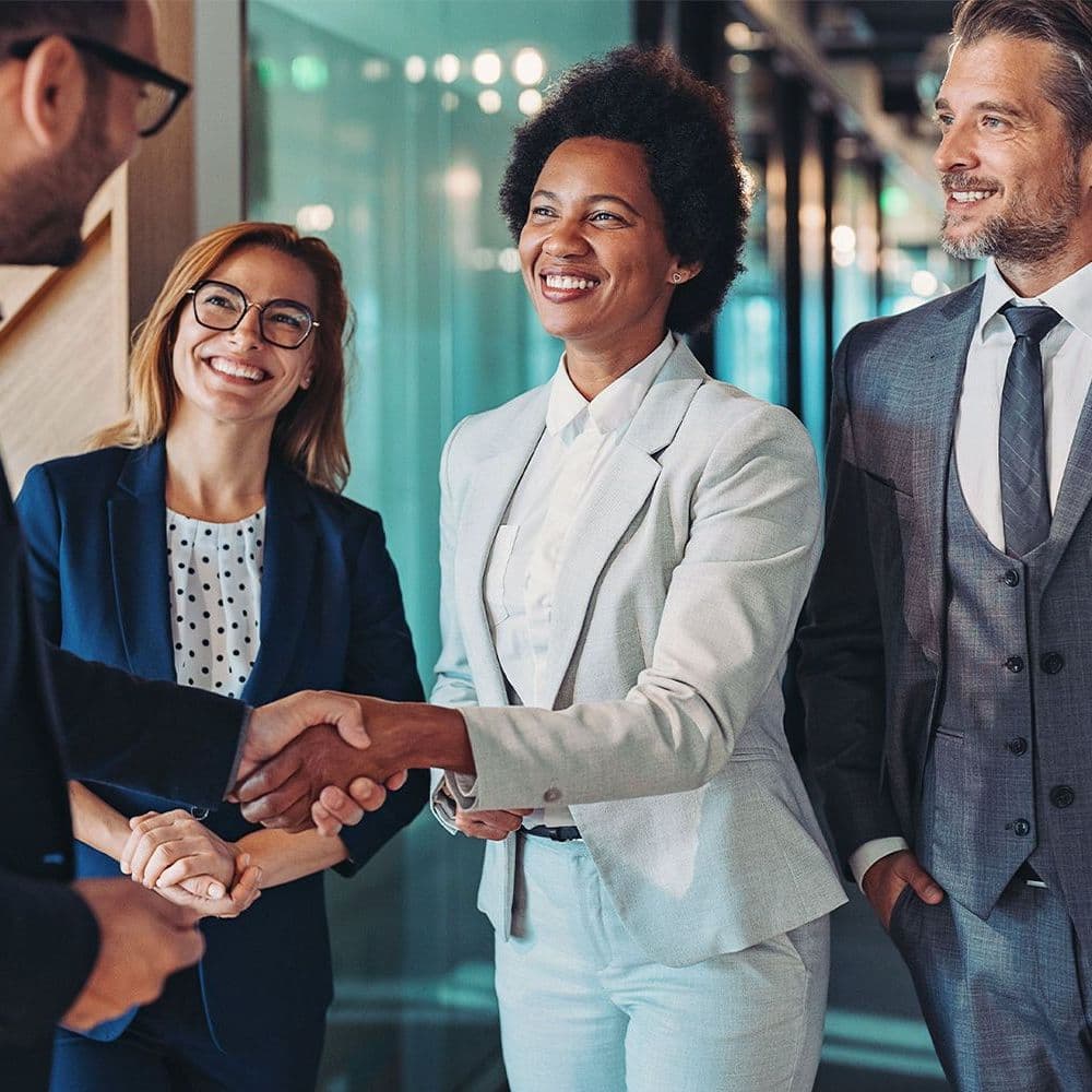 Vier Personen in Businesskleidung begrüßen sich freundlich in einem modernen Büroflur mit Glaswänden und Holztreppe im Hintergrund
