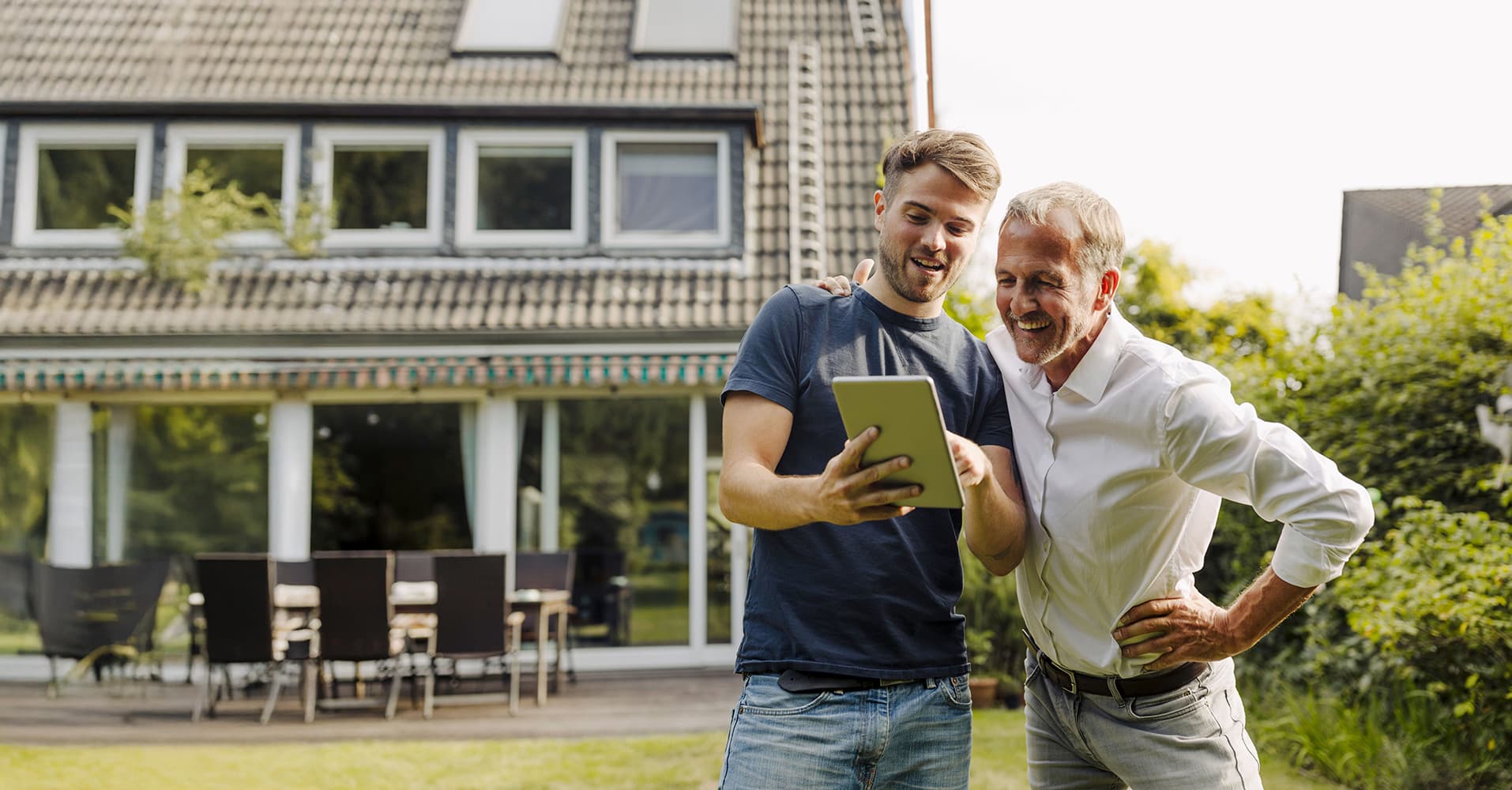 Zwei Männer stehen im Garten vor einem Einfamilienhaus und schauen gemeinsam auf ein Tablet