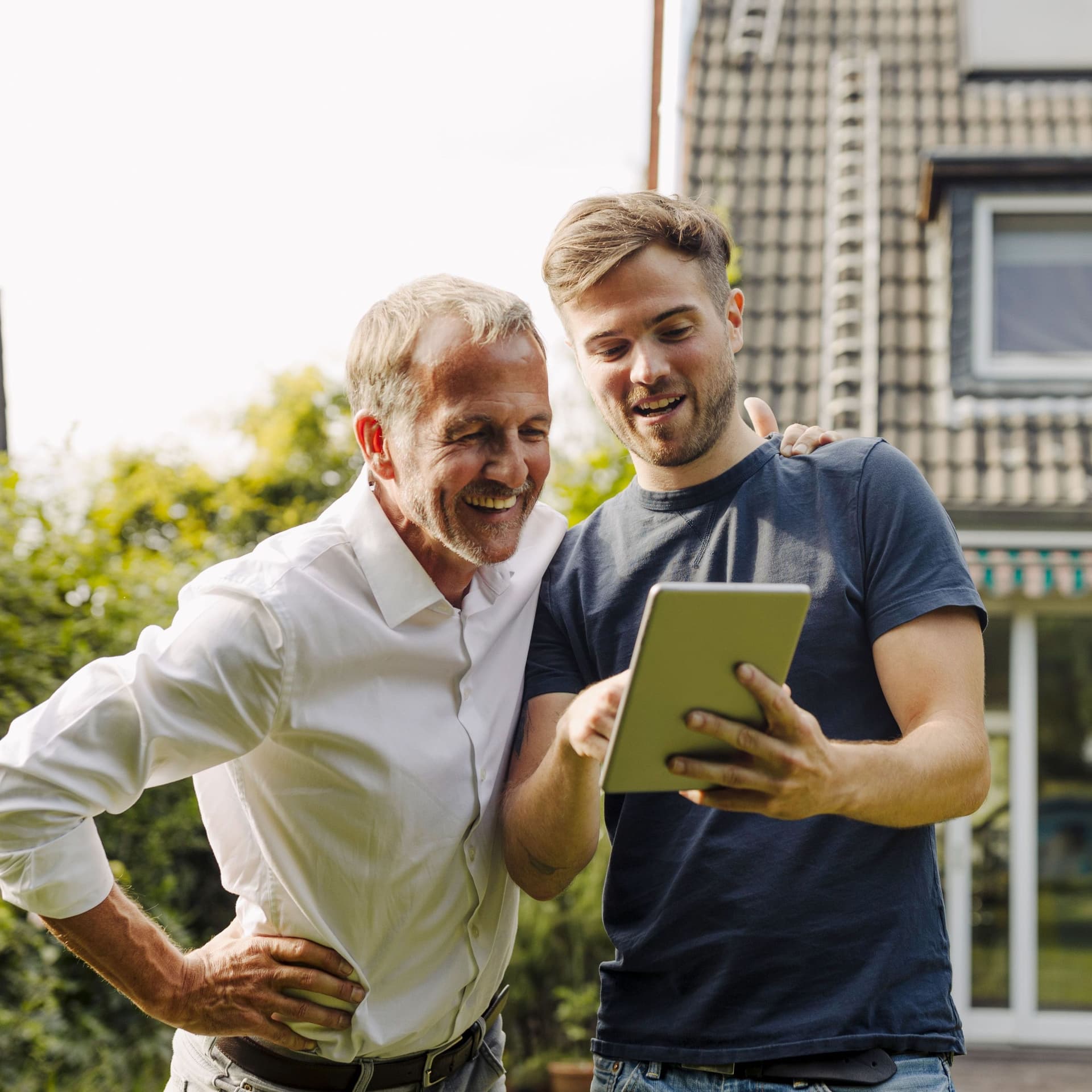 Vater und Sohn stehen in einem Garten vor einem Einfamilienhaus, der Sohn zeigt dem Vater etwas auf seinem Tablet