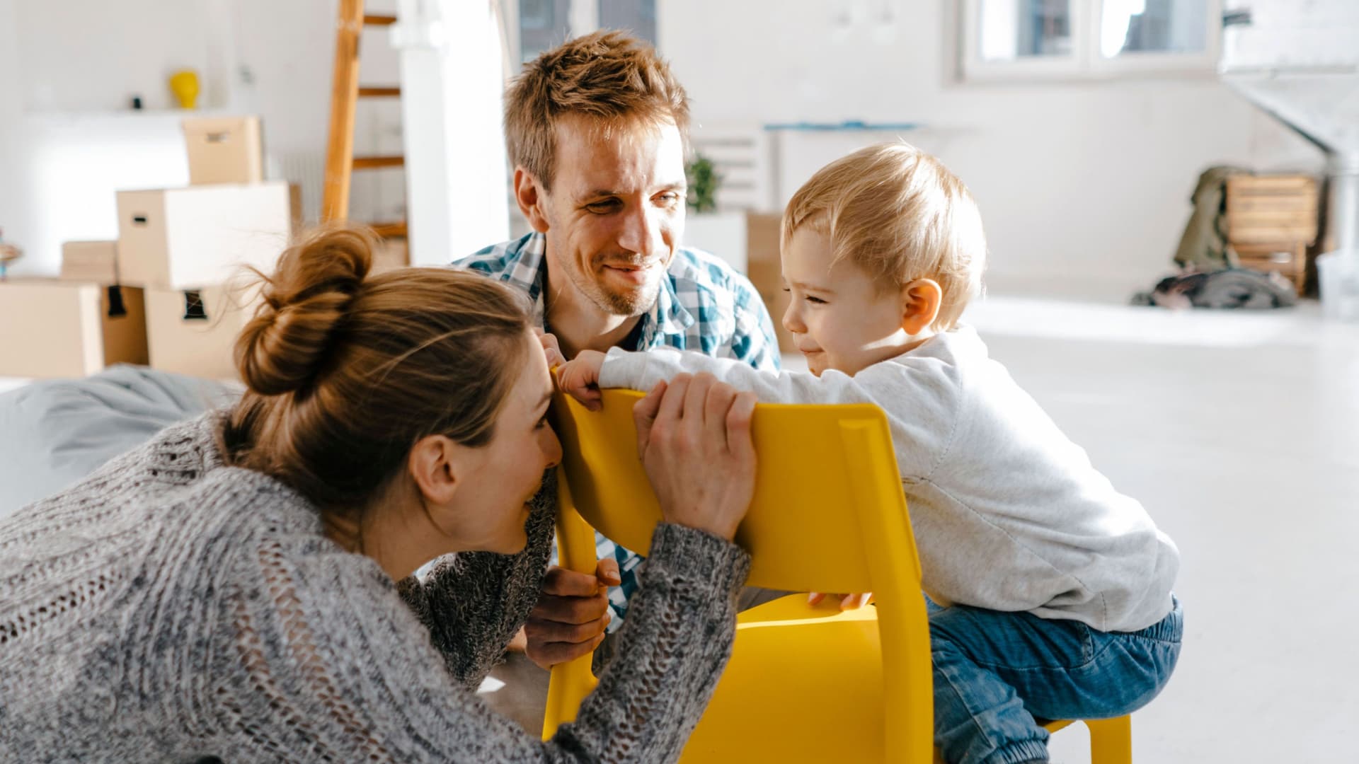 Junge Familie in einem modernen noch nicht fertig eingerichteten Raum. Die Mutter hockt vor einem gelben Stuhl und versteckt ihr Gesicht vor dem Kleinkind, was auf dem Stuhl sitzt.