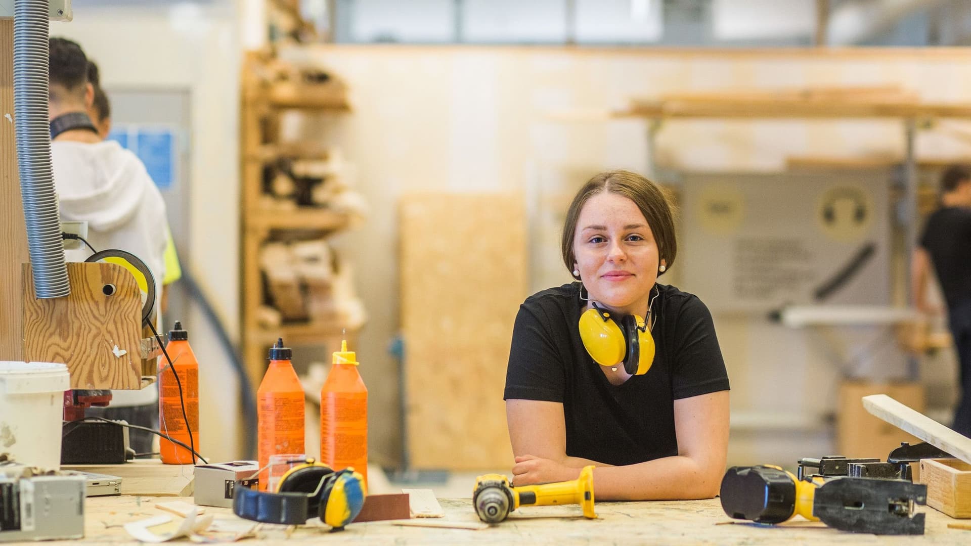 Eine junge Frau lehnt auf dem Tisch in einer Schreinerei. Sie lächelt in die Kamera. Um den Hals trägt sie einen Gehörschutz.