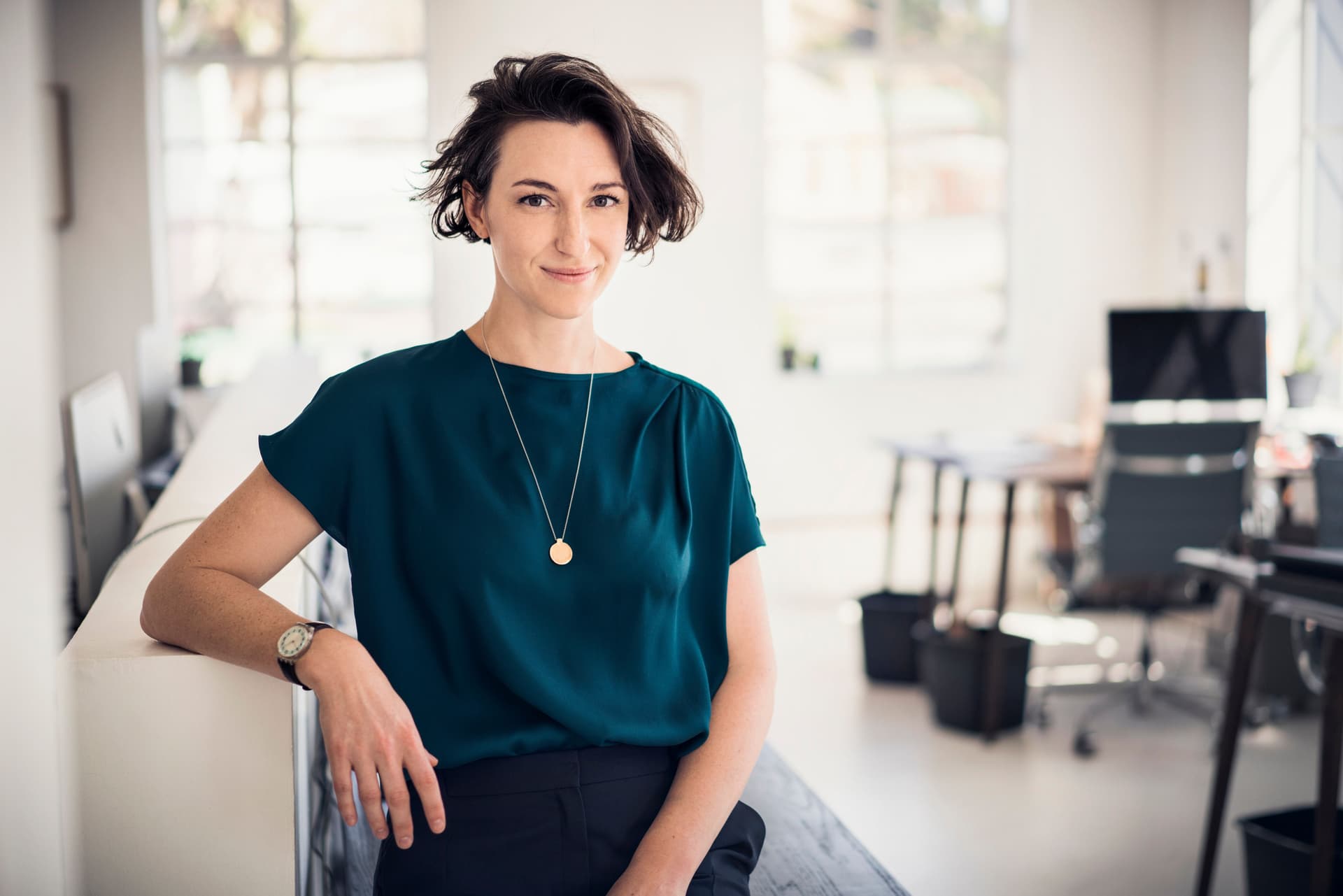Ein Foto einer lächelnden Frau, die auf einem Tisch in einem hellen, modernen Büro sitzt.