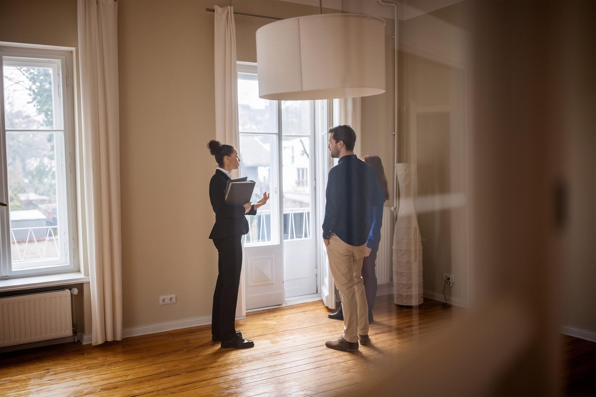 Immobilienmaklerin steht mit einem jungen Paar in einem leeren Zimmer. Sie trägt einen Hosenanzug und hat eine Mappe in der Hand.