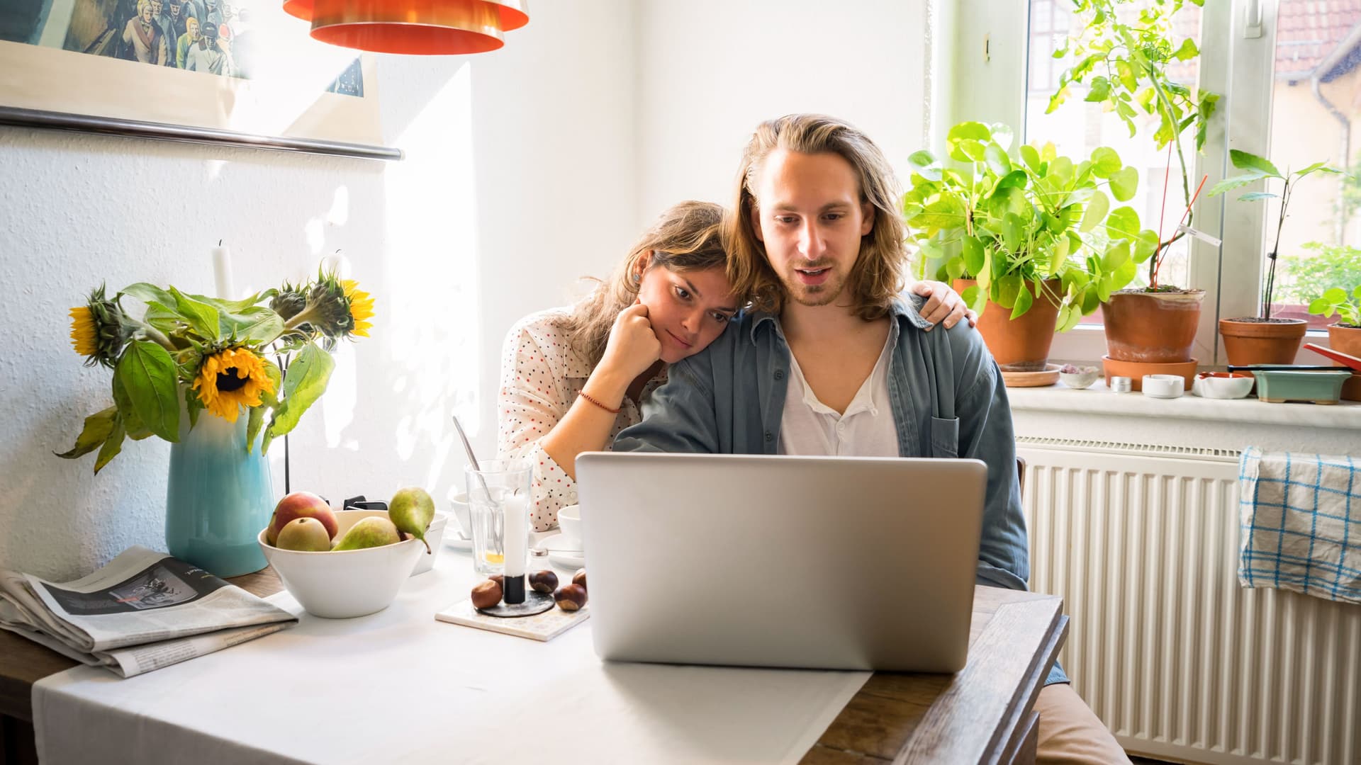 Ein junges Paar sitzt an einem Küchentisch vor einem Laptop. Die Frau hat den Kopf auf die Schulter des Mannes gelegt.