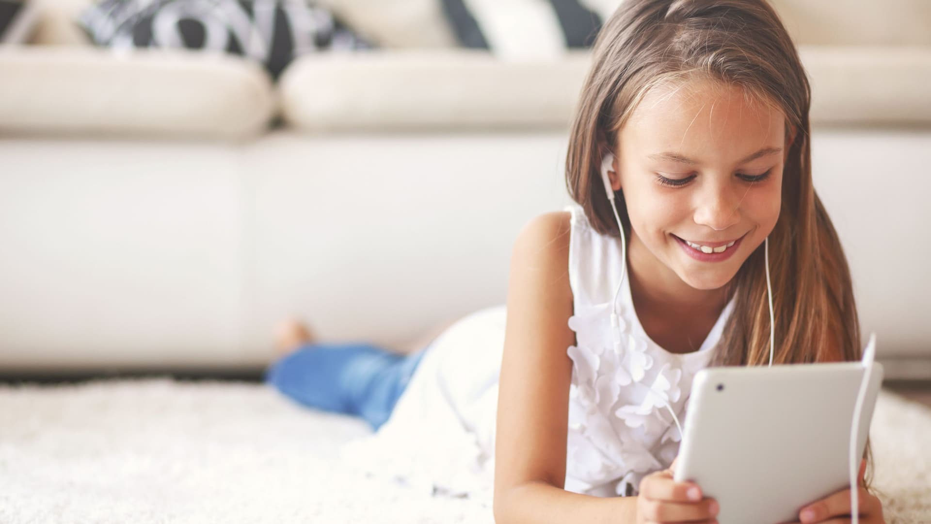 sicher im Internet surfen Mädchen im Grundschulalter liegt auf weißem Teppich mit Tablet in den Händen. Sie lächelt und hat Kopfhörer im Ohr.