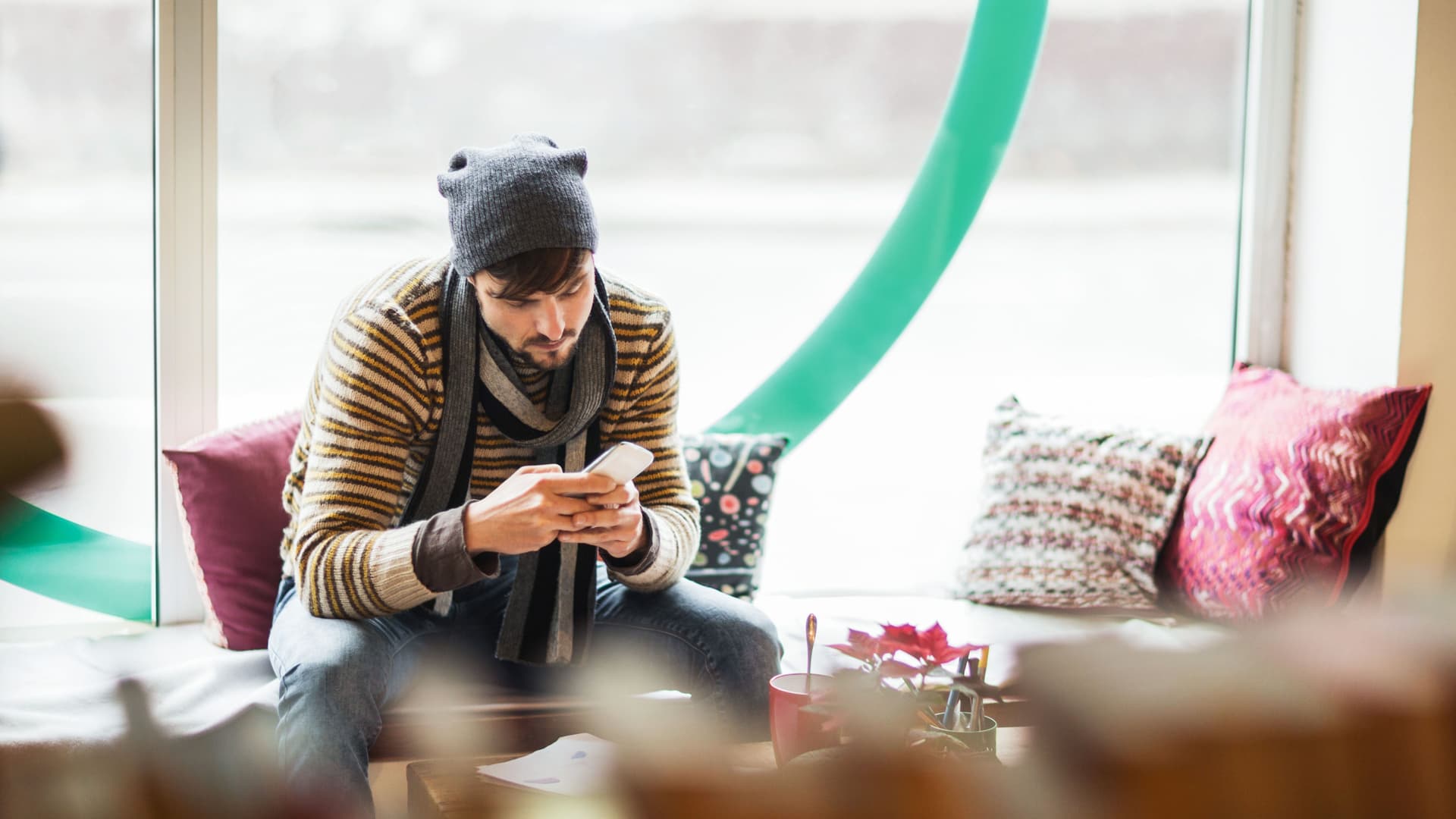 Junger Mann mit Mütze und Schal im Cafe schaut auf sein Handy.