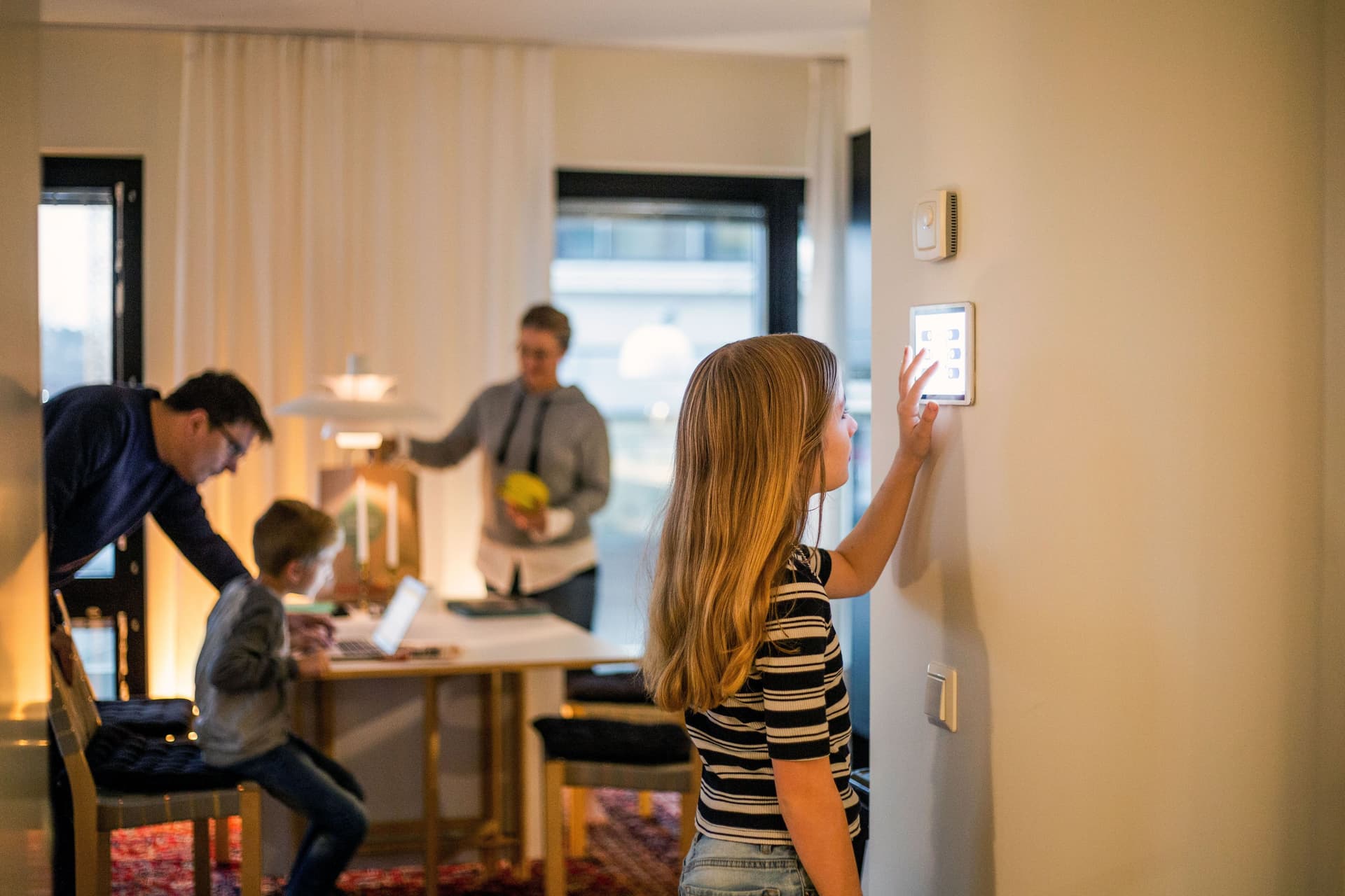 Mädchen bedient digitales Smart Home Display an der Wand. Im Hintergrund ist unscharf die Familie zu erkennen.