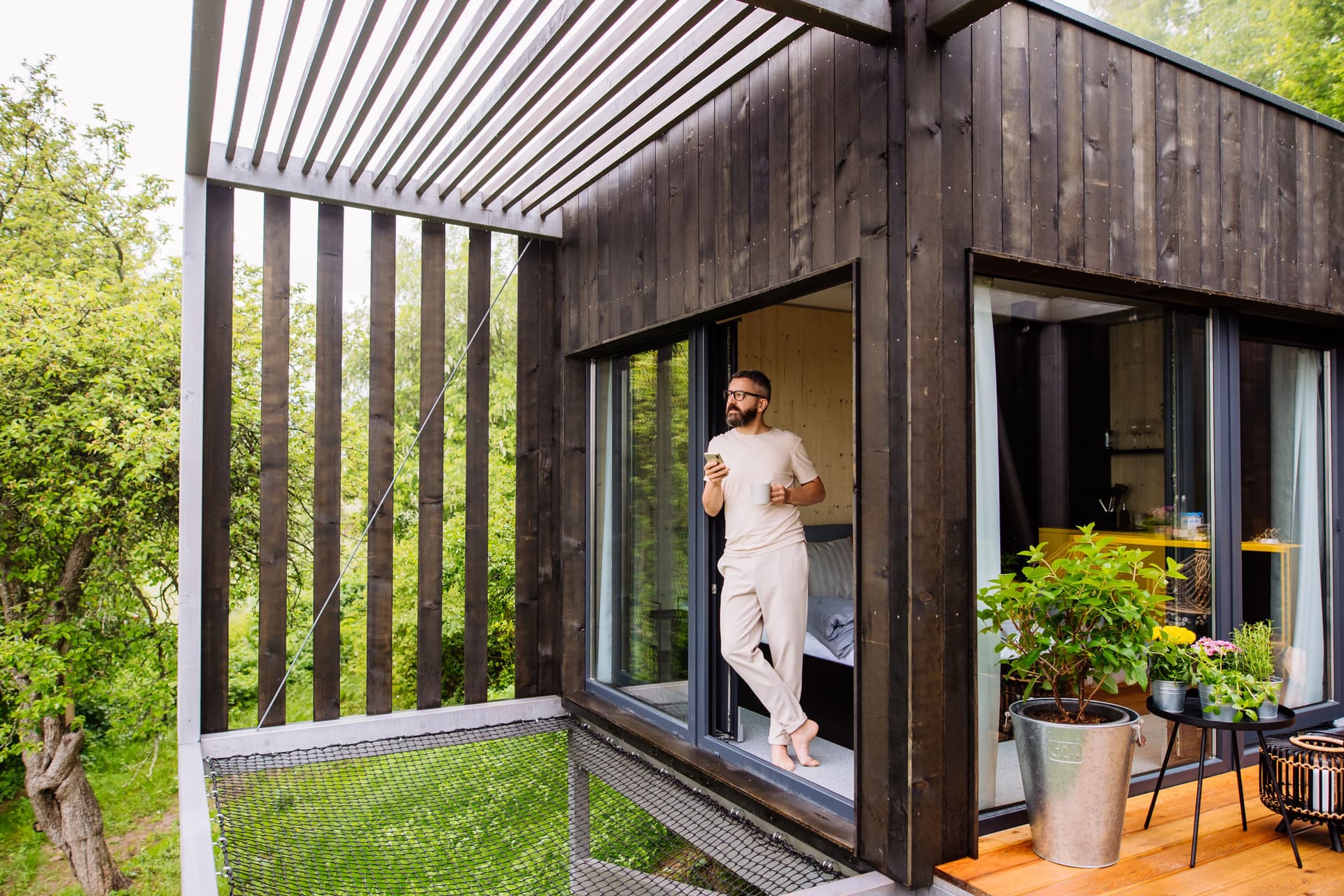 Ein Mann steht barfuß mit einer Tasse Kaffee und einem Smartphone am Fenster eines Tiny Houses und blickt dabei in die Natur.