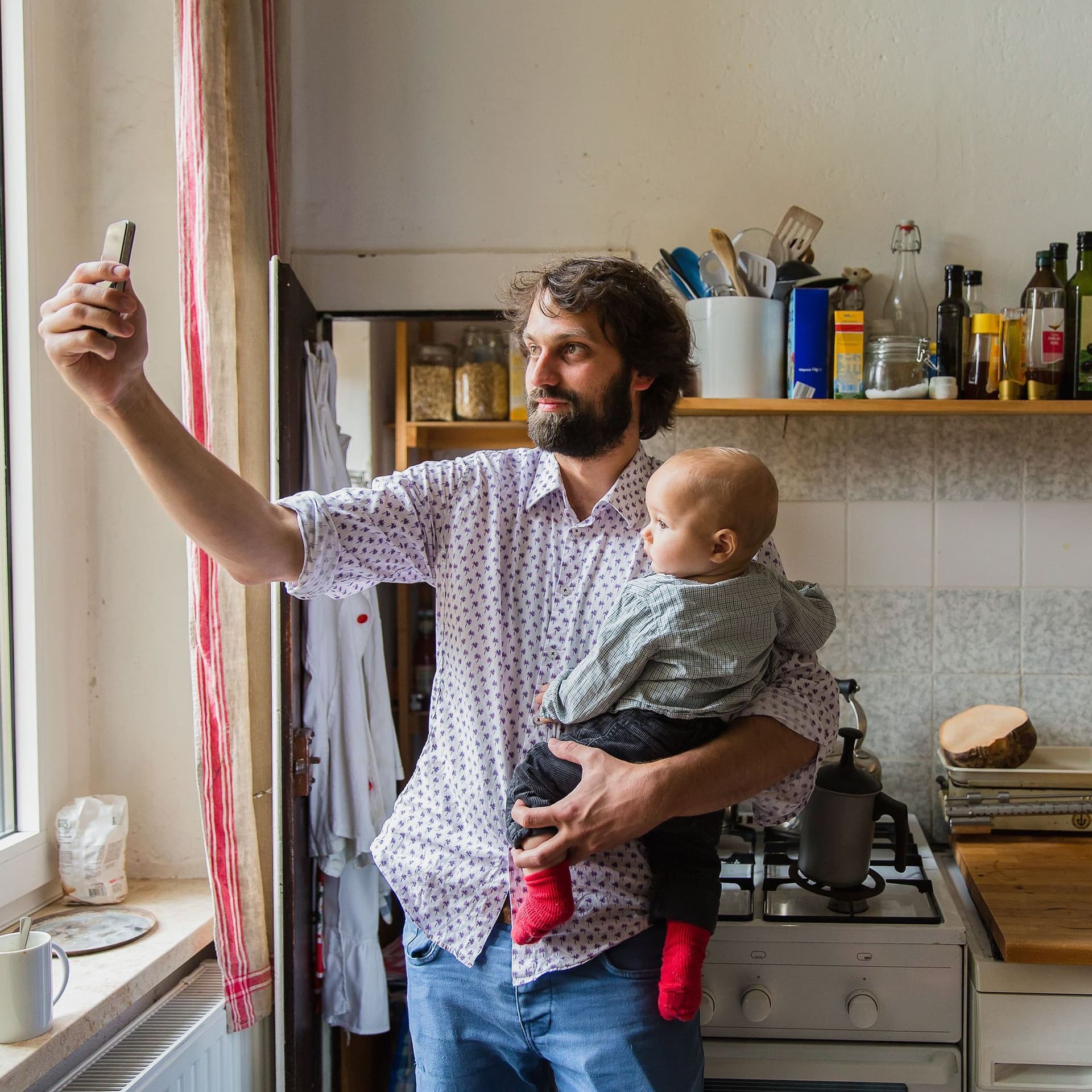 Ein junger Vater mit Baby auf dem Arm steht in einer Küche und macht ein Selfie von sich und seinem Kind.