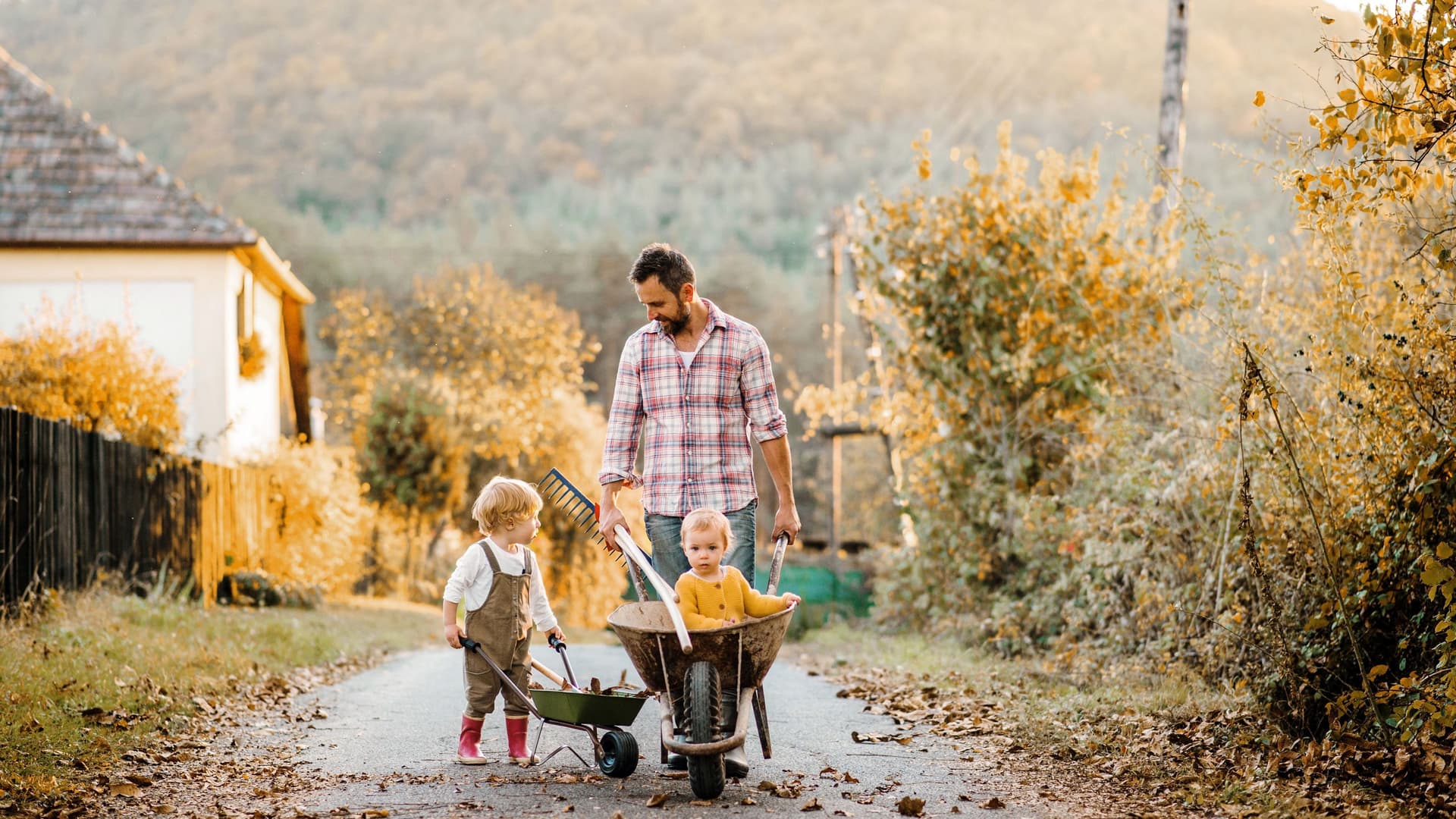 Ein Vater läuft mit zwei Kleinkindern und Schubkarren auf einem ländlichen Weg.