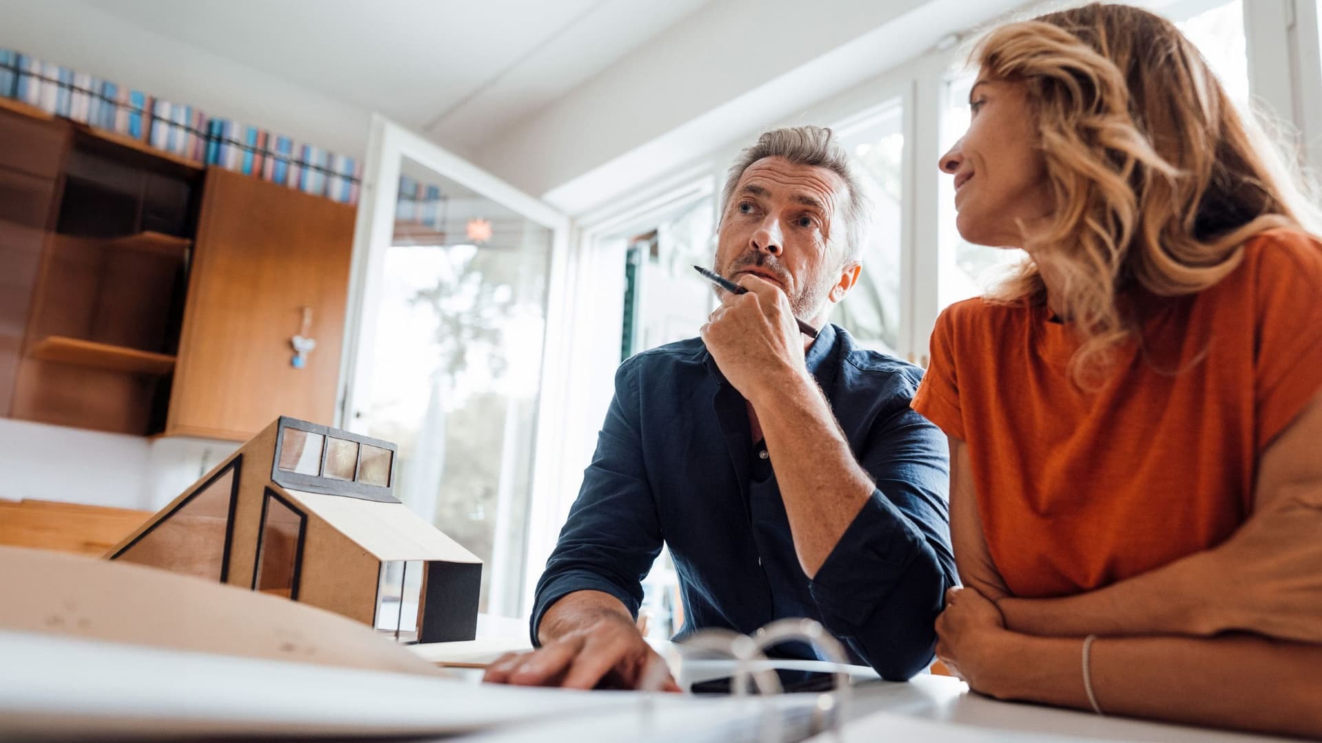 Mann und Frau sitzen nachdenklich an einem Tisch mit Hausmodell und Bauplänen