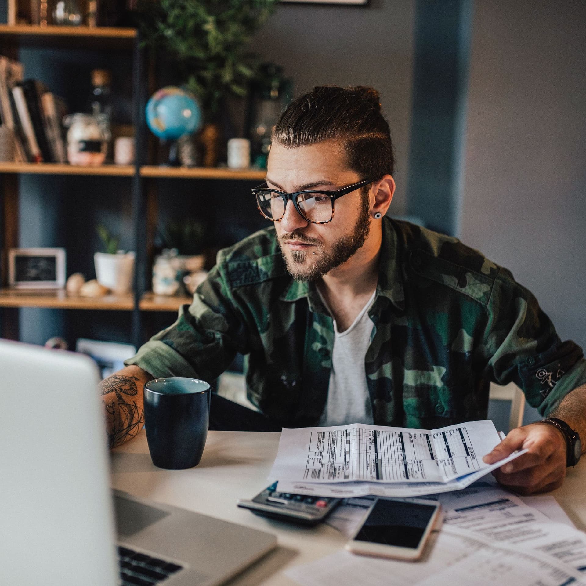 Blick über einen Laptop auf einen jungen Mann mit Unterlagen und Taschenrechner vor sich.