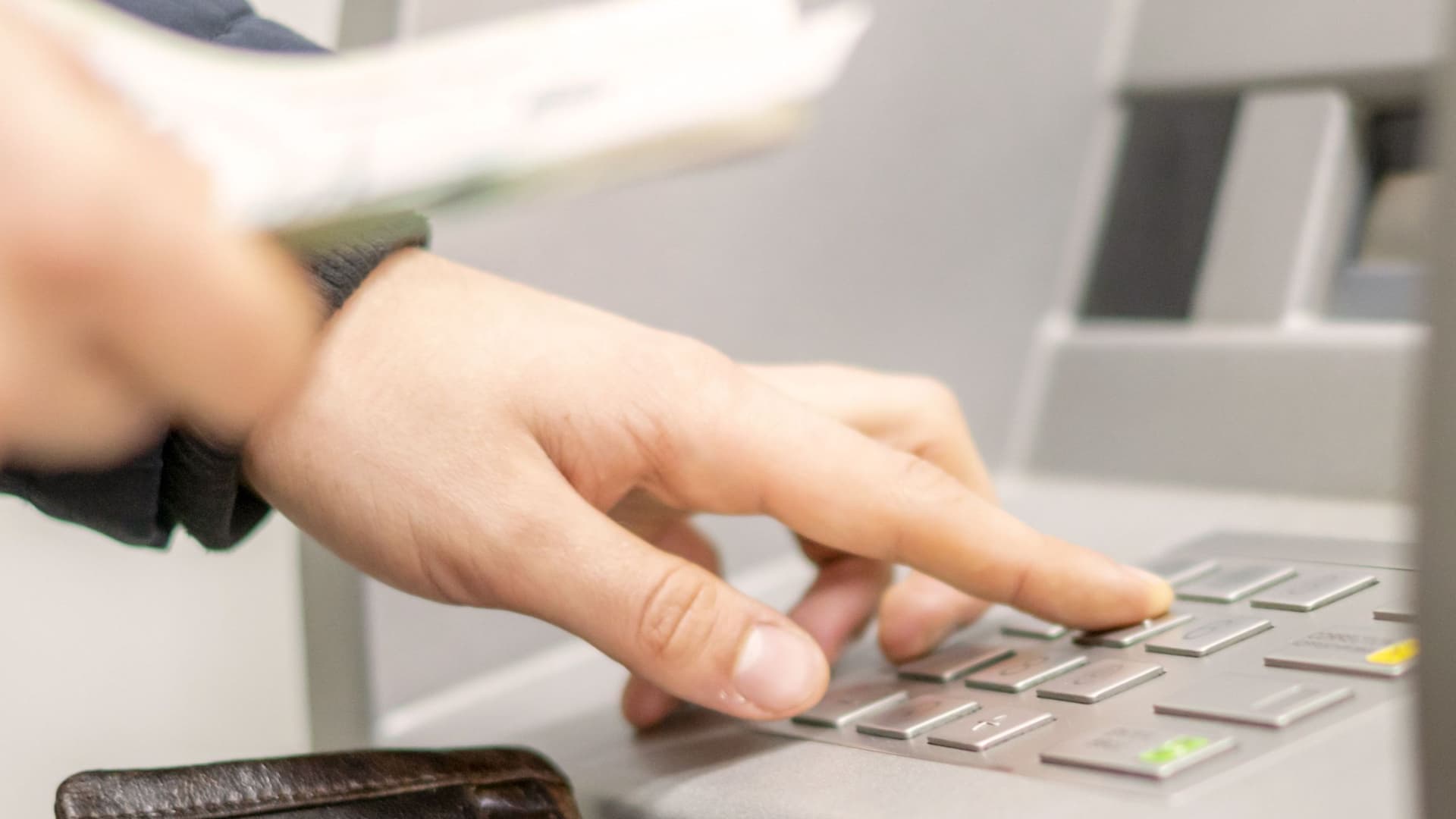 Person mit Geldscheinen in der Hand tippt an einem Geldautomaten die Geheimzahl ein.
