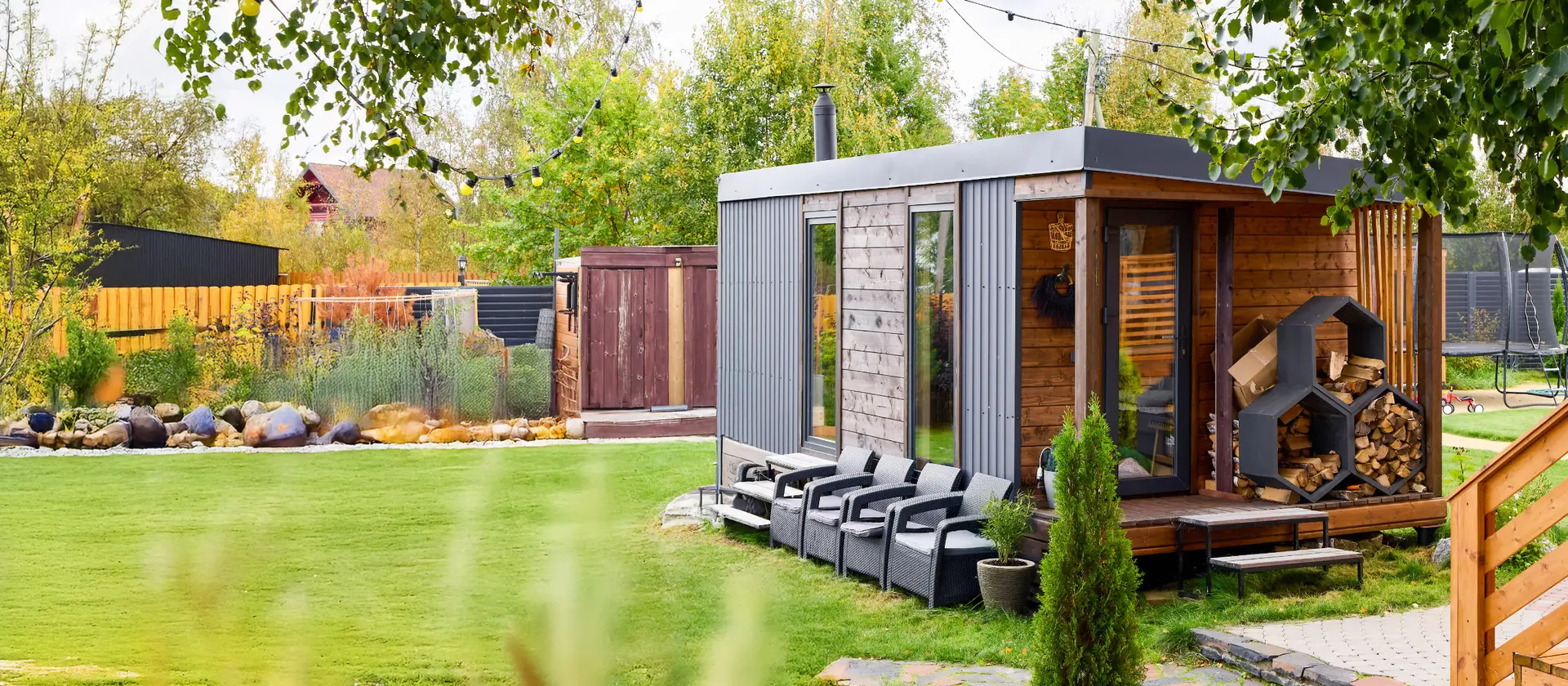 Kleiner moderner Gartensauna‑Container aus Holz mit Holzstapel, Terrassenstühlen und Rasen im Vordergrund.