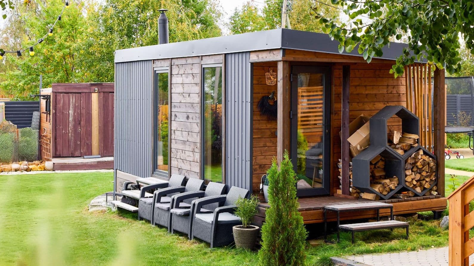 Kleiner moderner Gartensauna‑Container aus Holz mit Holzstapel, Terrassenstühlen und Rasen im Vordergrund.