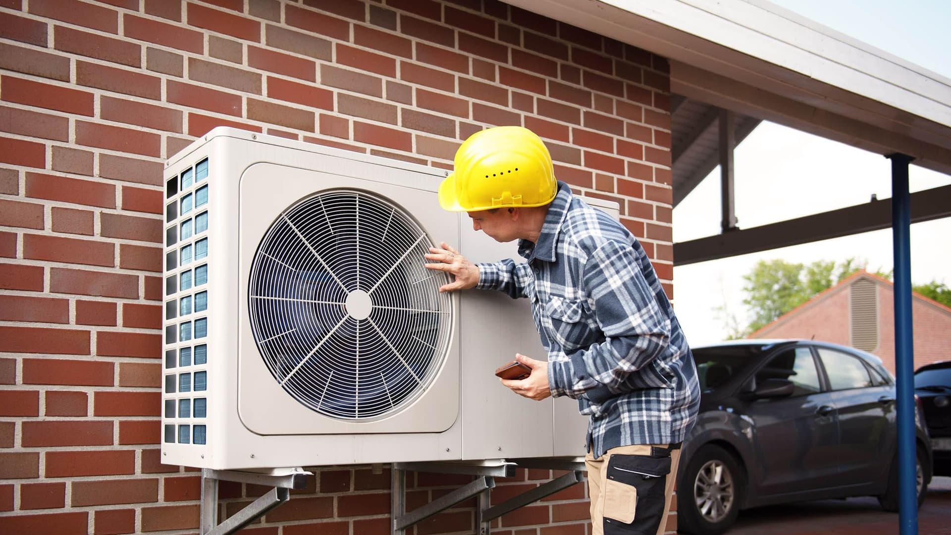 Ein Heizungsbauer in Arbeitskleidung prüft die Funktion einer Luft-Wärmepumpe im Außenbereich eines Hauses.