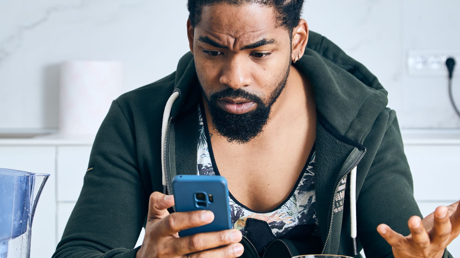 Ein Mann mit Bart sitzt in einer Küche und schaut mit verwirrtem, besorgtem Gesichtsausdruck auf sein Smartphone, während er eine Hand fragend hebt.