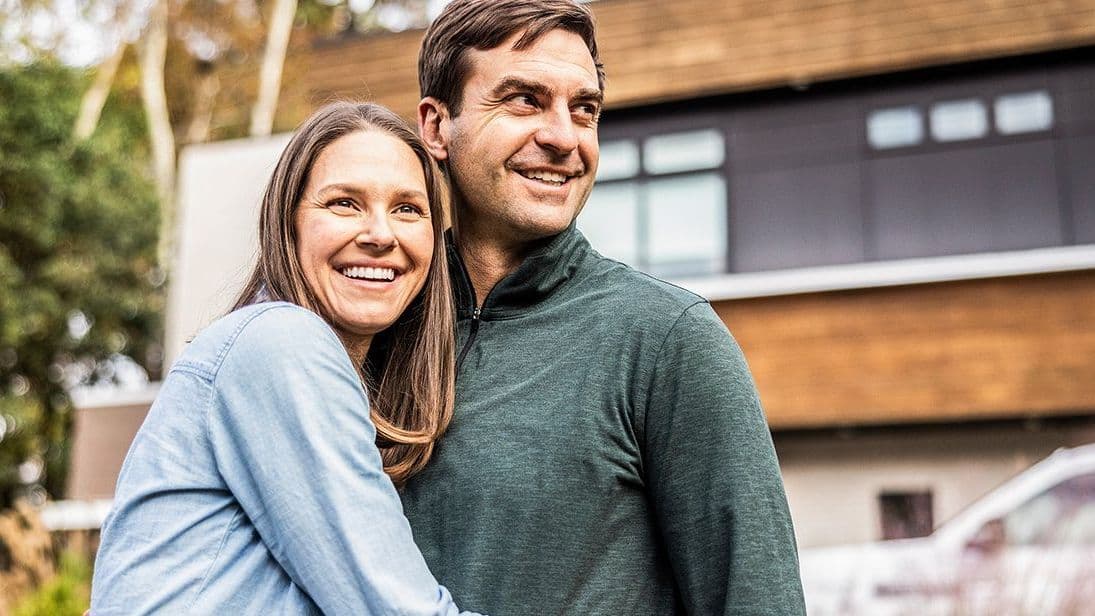 Ein lächelndes Paar umarmt sich vor einem modernen Einfamilienhaus. Beide wirken glücklich und blicken optimistisch nach vorne. Im Hintergrund sind Fassade, Fenster und ein geparktes Auto zu sehen.