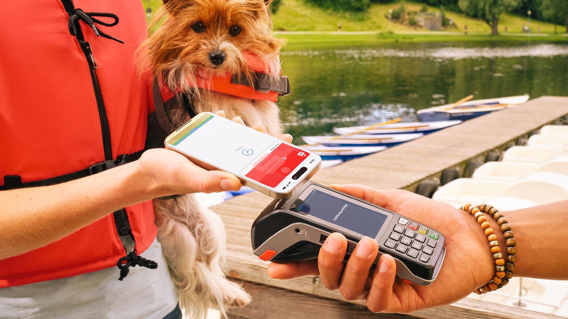 Eine Person in Schwimmweste hält ein Smartphone mit geöffneter Bezahlfunktion an ein Kartenterminal, während sie einen kleinen Hund am Seeufer trägt.