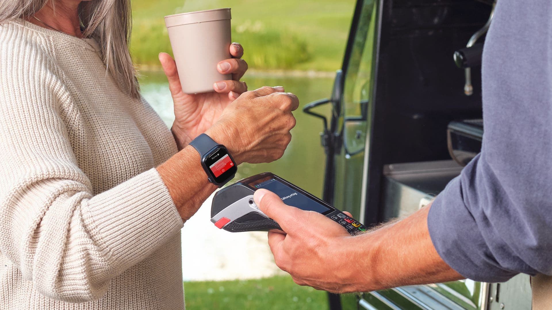 Eine Frau hält einen Kaffeebecher in der Hand und bezahlt mit ihrer Smartwatch kontaktlos an einem mobilen Kartenterminal vor einem Coffee-Truck.