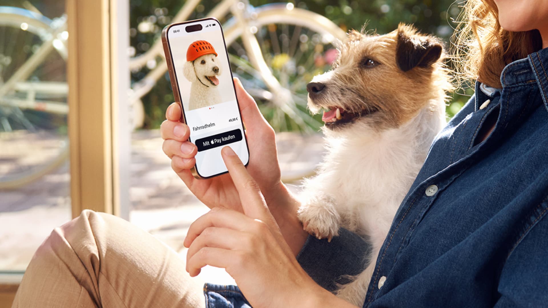 Eine Frau ist sitzend im Anschnitt zu sehen und hat einen Hund auf dem Schoß und nutzt ihr Smartphone, um online einen Fahrradhelm für Hunde zu kaufen. Im Hintergrund ist ein sonniger Garten mit einem Fahrrad zu sehen.