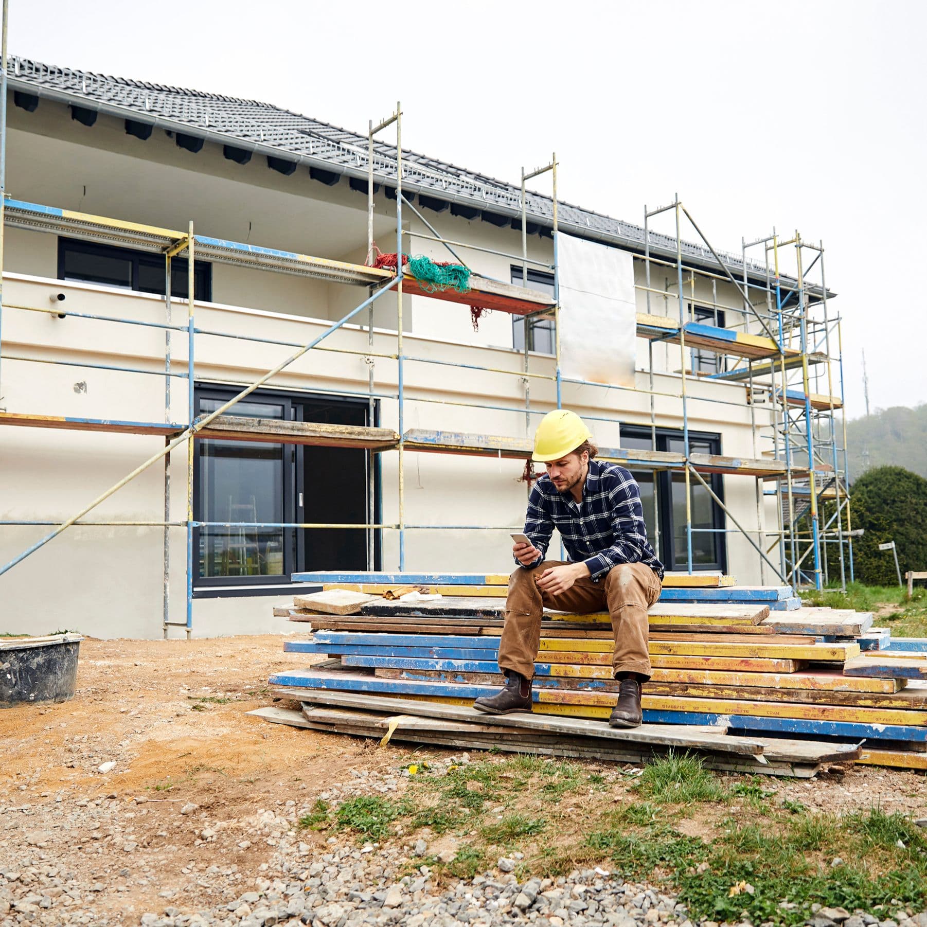 Ein Bauarbeiter mit Helm sitzt vor einer menschenleeren Baustelle auf einem Stapel Gerüstbrettern und schaut mit ernstem Gesichtsausdruck auf sein Smartphone.
