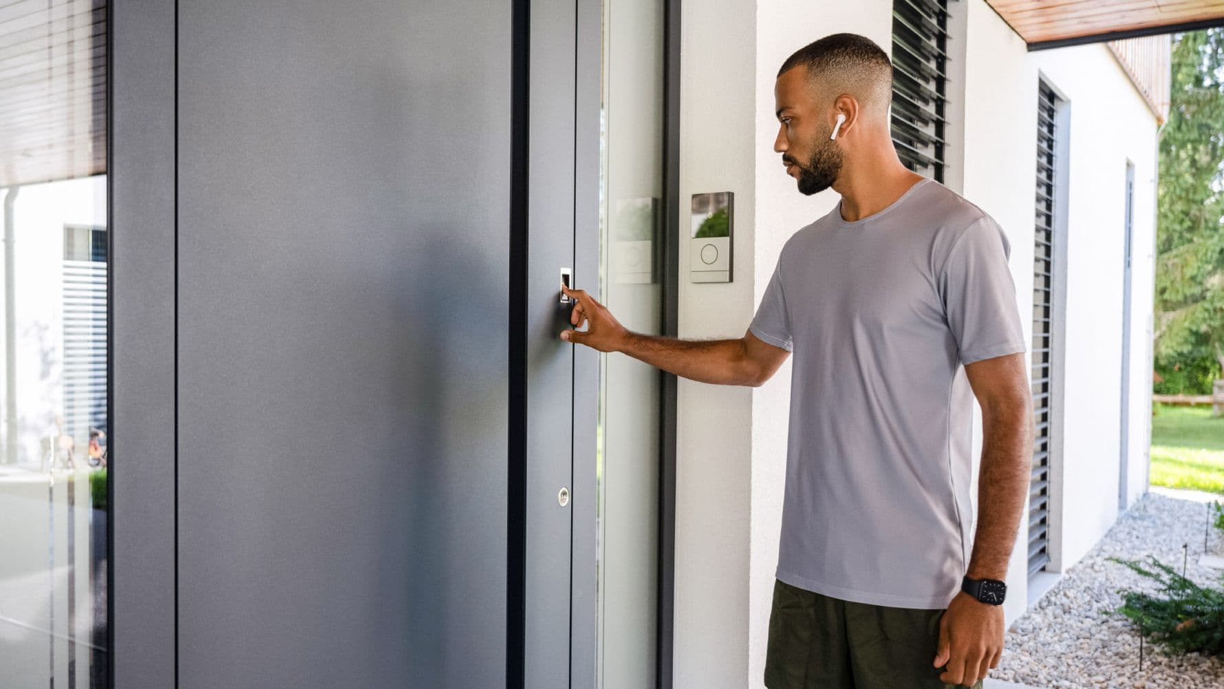 Ein Mann in sportlicher Kleidung steht vor einer modernen Haustür und greift zur Türklinke. Er trägt kabellose Ohrhörer und befindet sich vor einem hellen Wohnhaus.