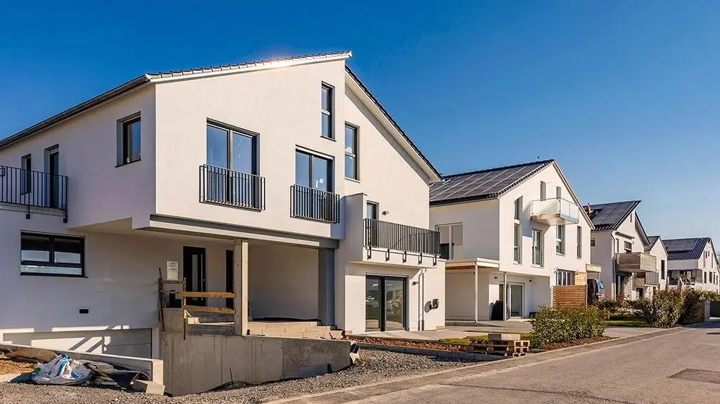 Eine Außenaufnahme eines Straßenzuges mit neugebauten Einfamilienhäusern. Ein weißes Haus ist zentral im Bild. Ähnliche Häuser reihen sich anhand einer Fluchtlinie hintereinander.