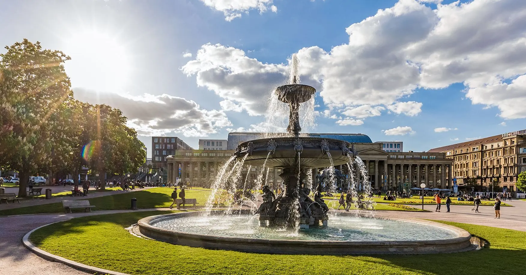 Öffentlicher Park in Stuttgart mit einem großen Springbrunnen im Vordergrund, Sonnenschein links, Menschen auf den Wiesen und klassizistischen Gebäuden im Hintergrund.
