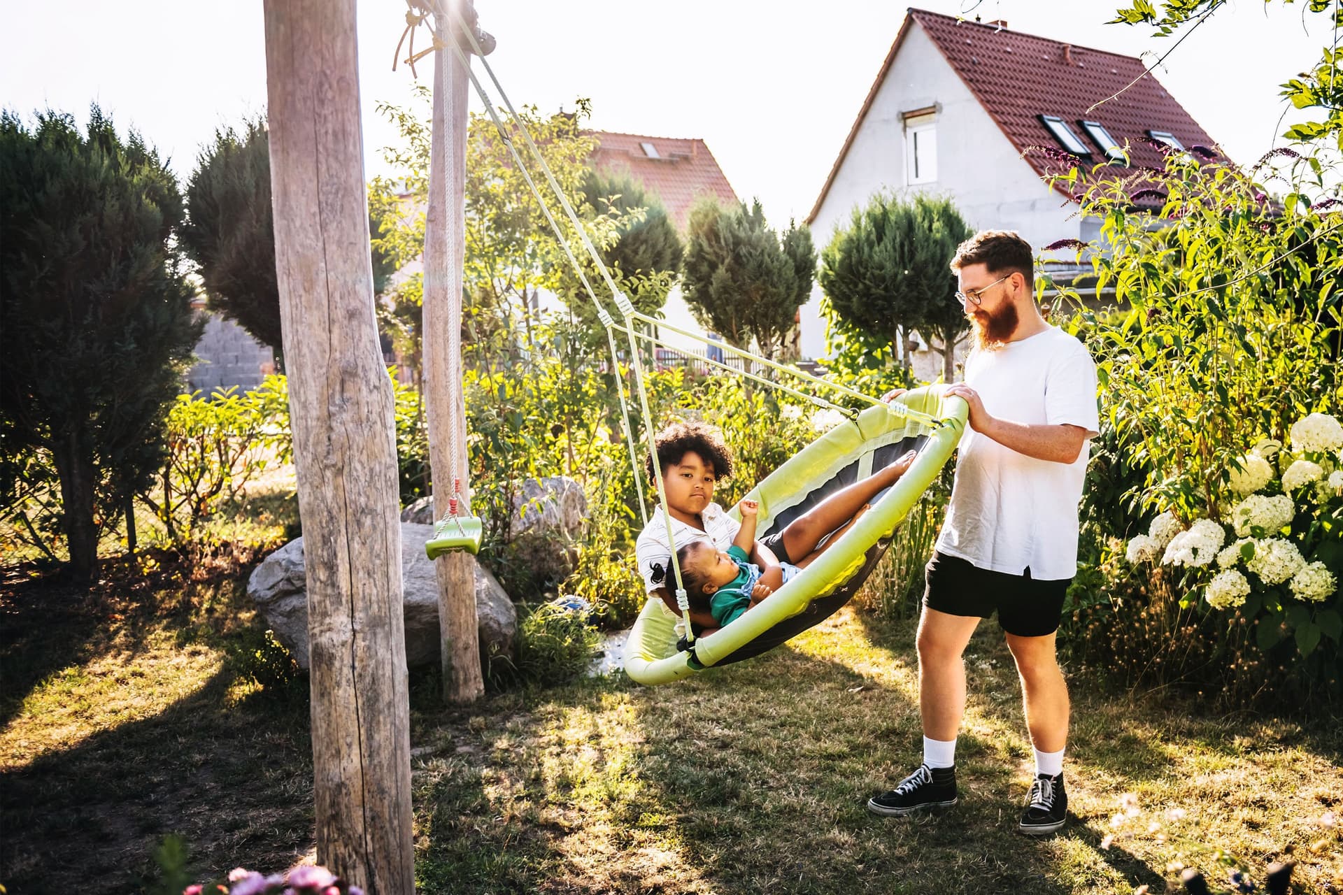 Ein Mann schubst eine große Schaukel mit zwei Kindern in einem grünen Garten an. Im Hintergrund sieht man Einfamilienhäuser.