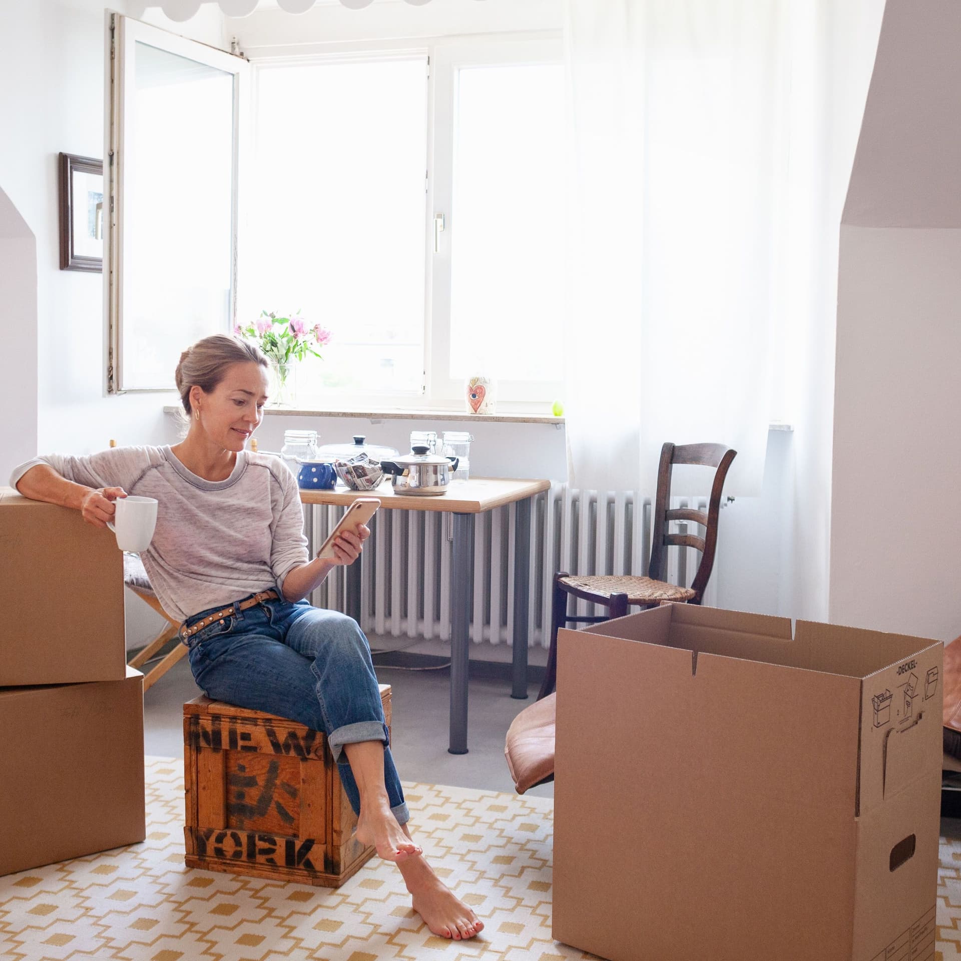 Frau sitzt auf gepackten Kisten in ihrer Wohnung. Sie schaut auf ihr Smartphone.