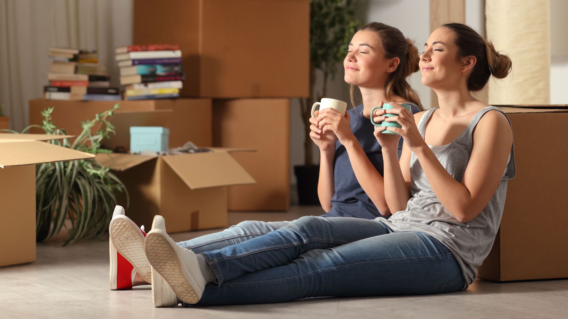 Zwei junge Frauen sitzen mit einer Tasse an einen Umzugskarton gelehnt und haben die Augen geschlossen.
