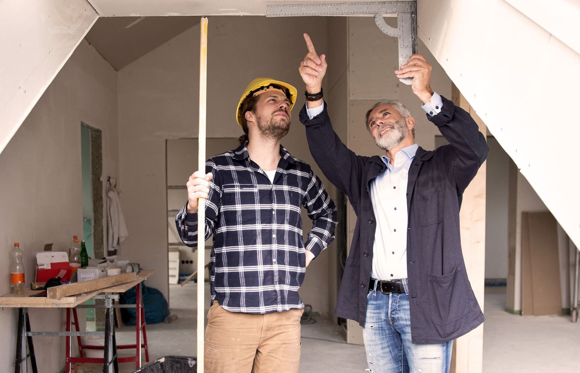 Zwei Männer arbeiten gemeinsam an einem Dachbodenausbau, während sie Materialien und Werkzeuge organisieren. Der Raum ist hell erleuchtet und zeigt eine Mischung aus Holzrahmen und Dämmmaterialien, die auf den bevorstehenden Umbau hinweisen.